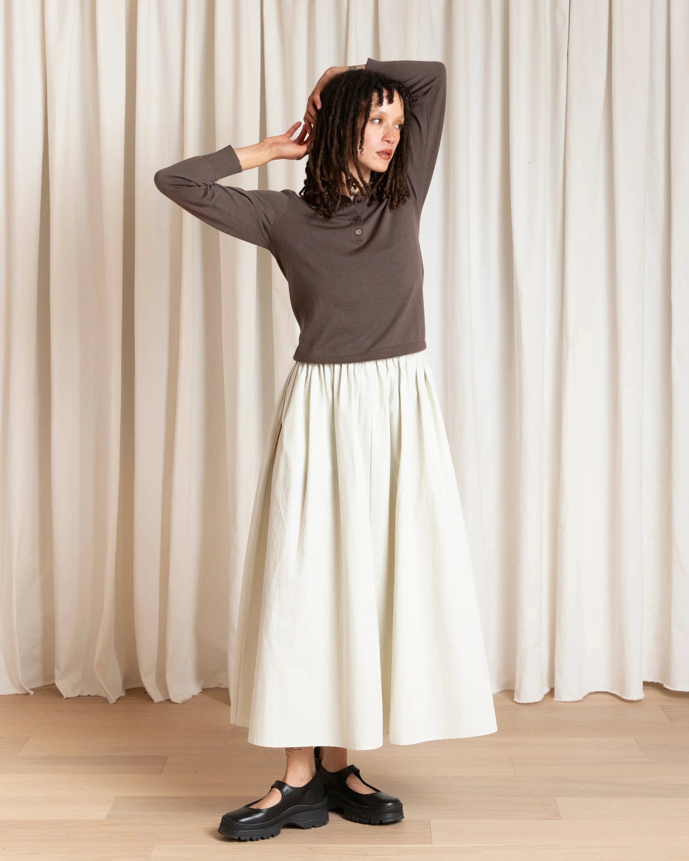 Person wearing a brown top and white skirt standing against a white curtain.