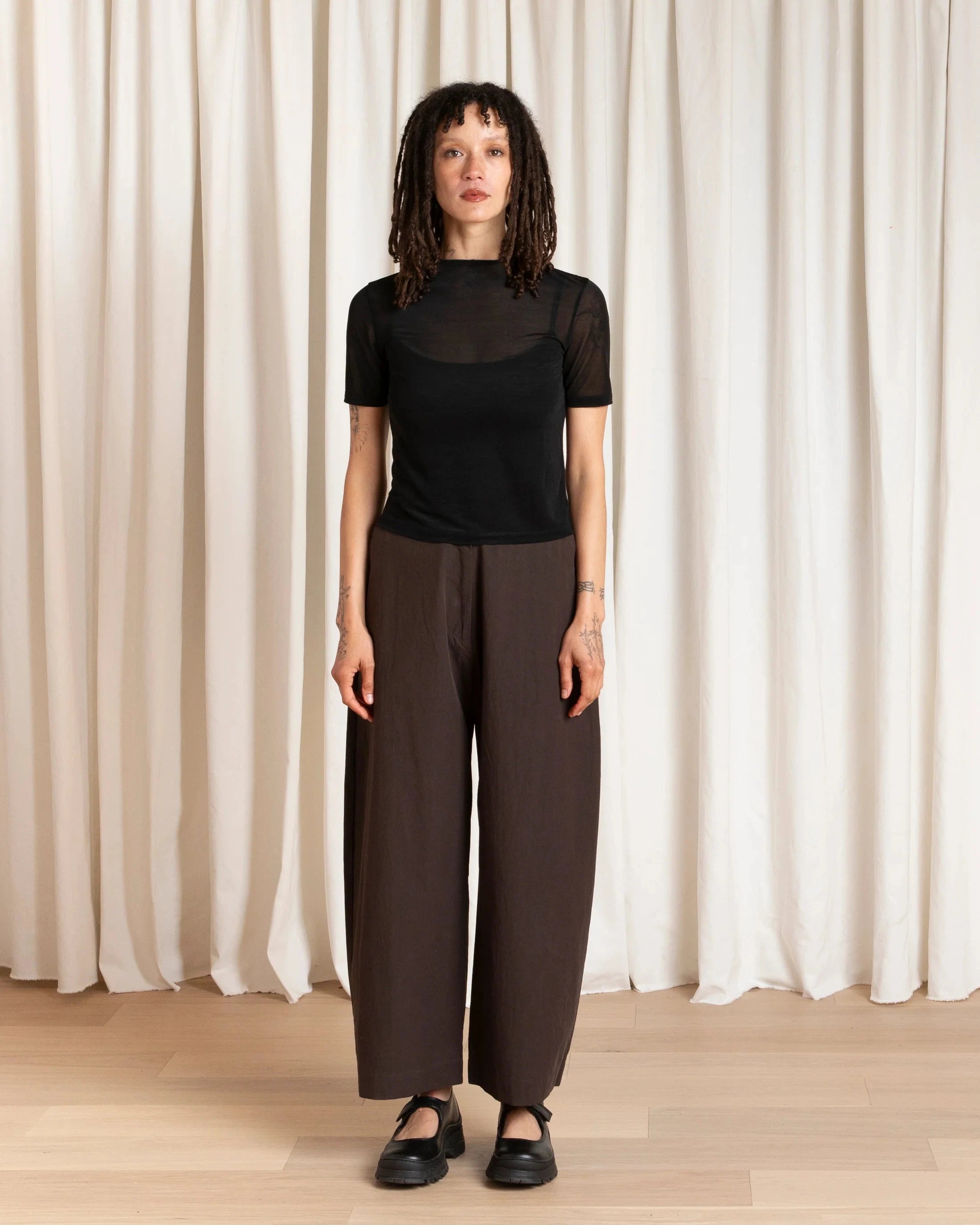 Person wearing a black top and brown pants standing against a white curtain.