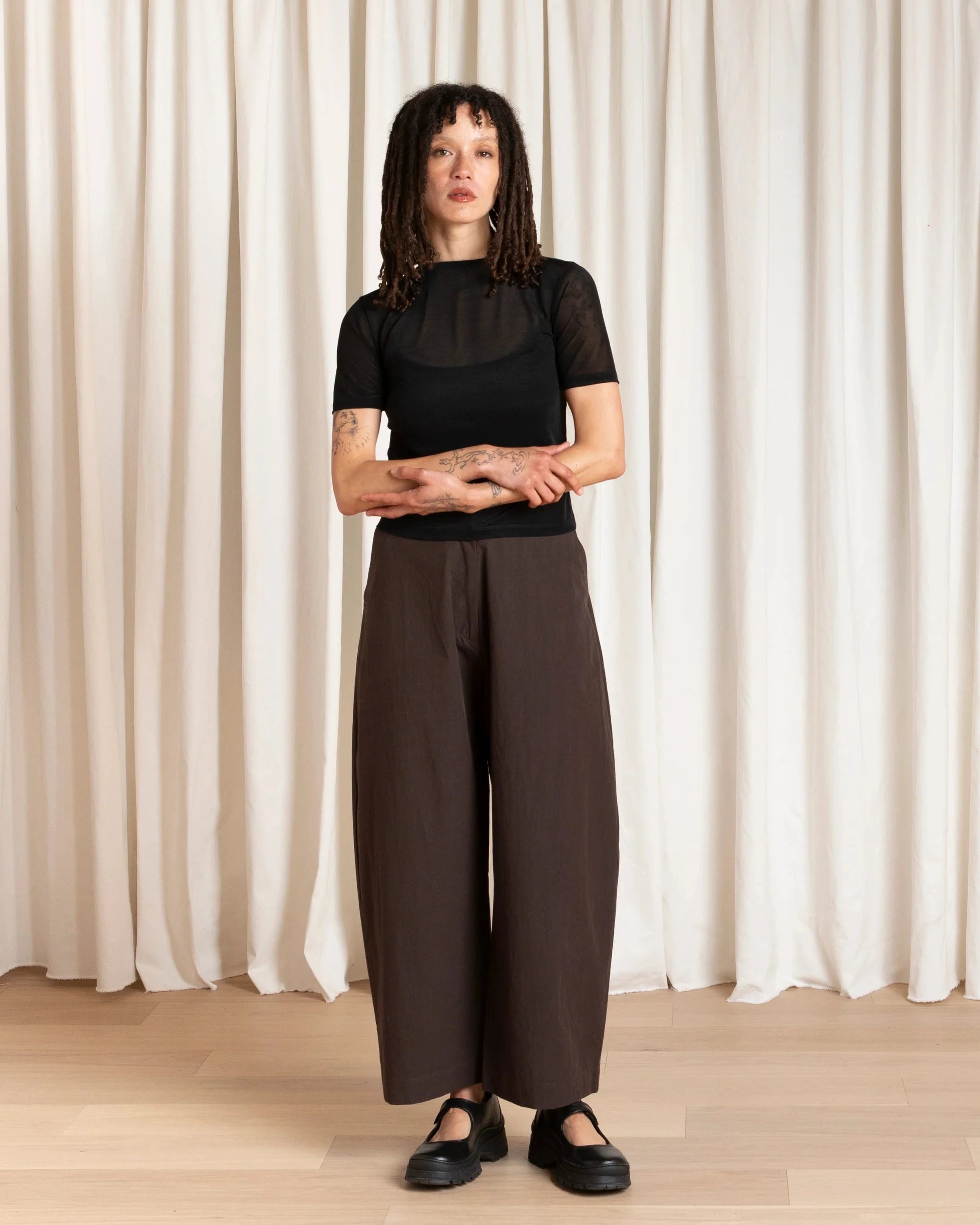 Person wearing a black top and brown pants standing against a white curtain.
