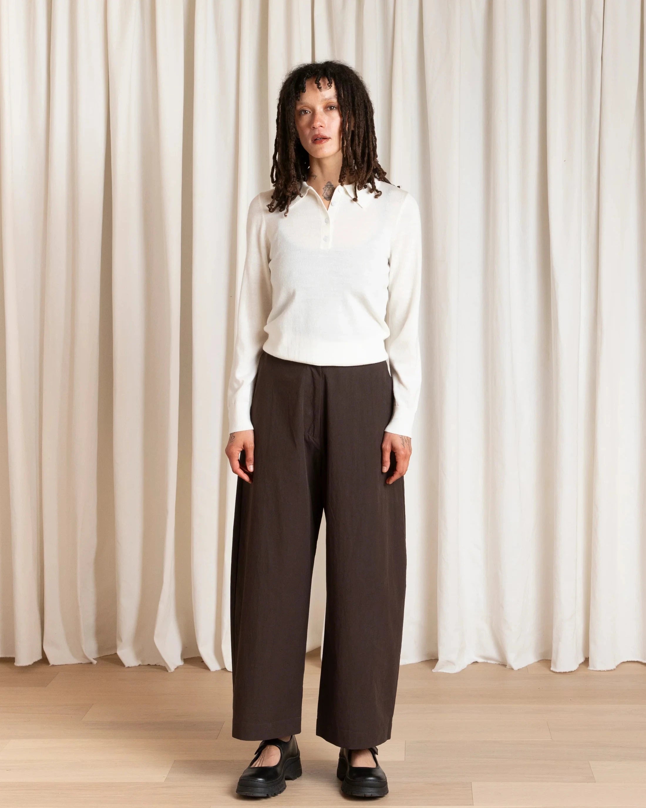 Woman wearing a white blouse and dark pants standing against a white curtain.