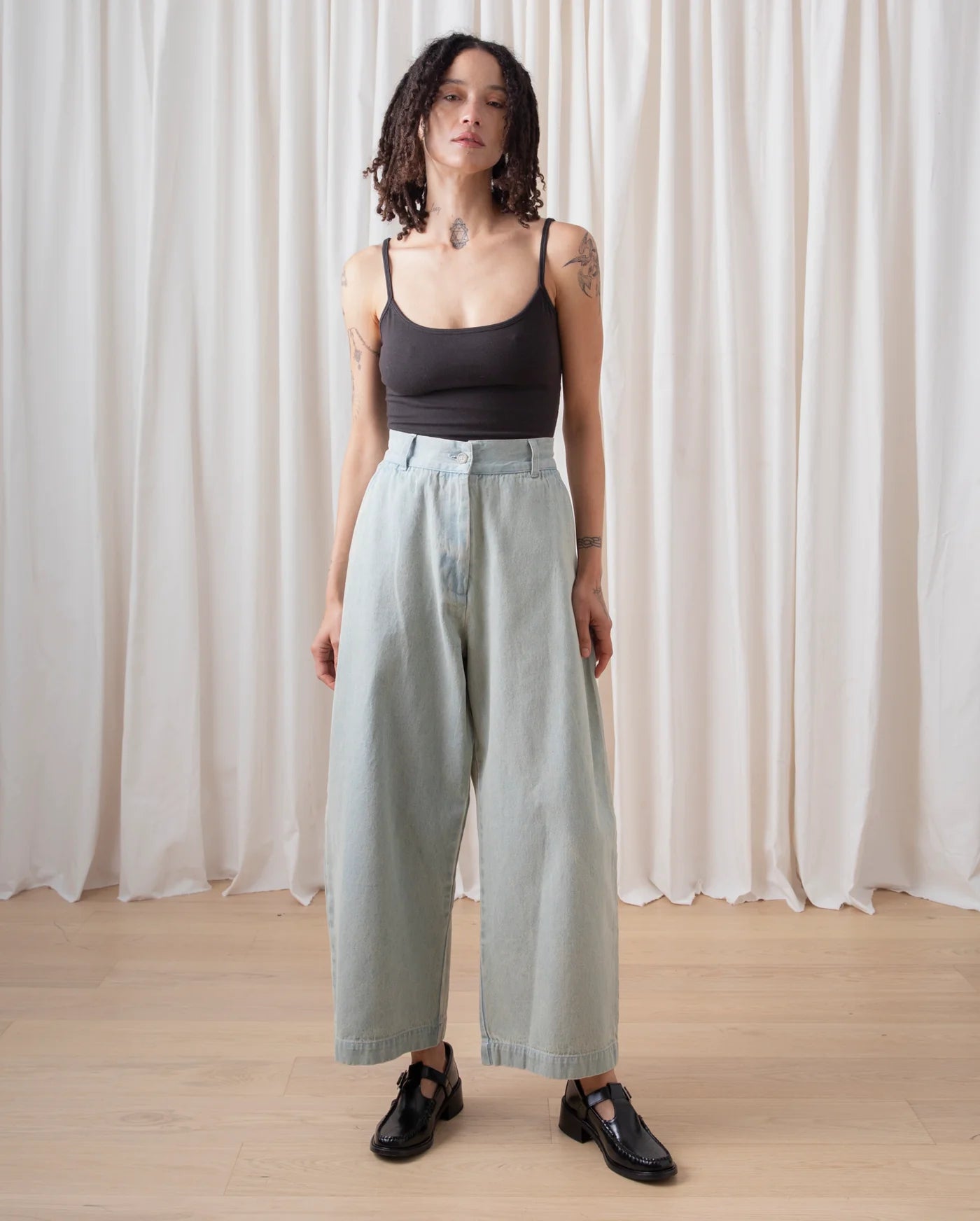 Person wearing a black tank top and light blue pants standing against a white curtain background