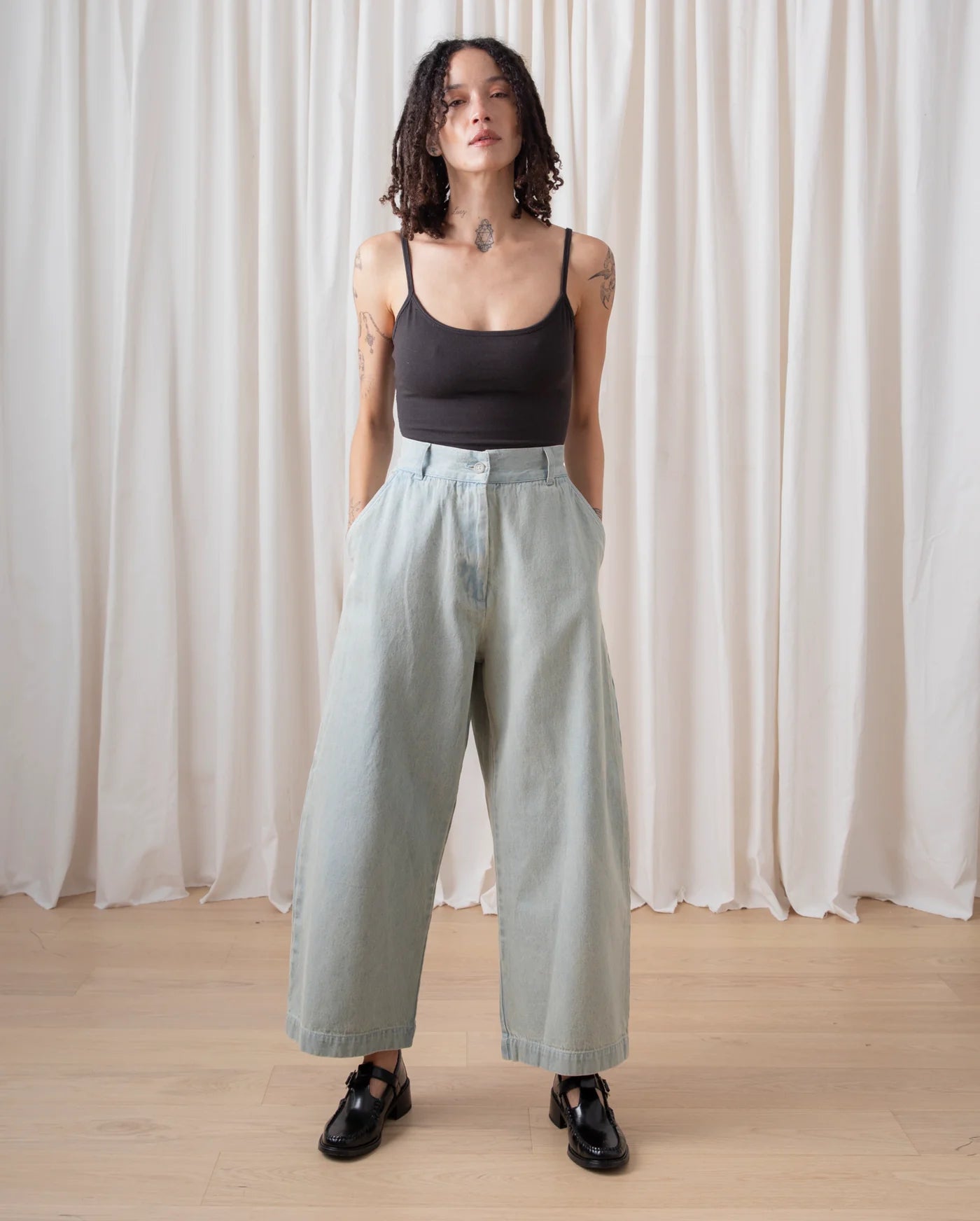 Person wearing a black tank top and light green pants standing against a white curtain.