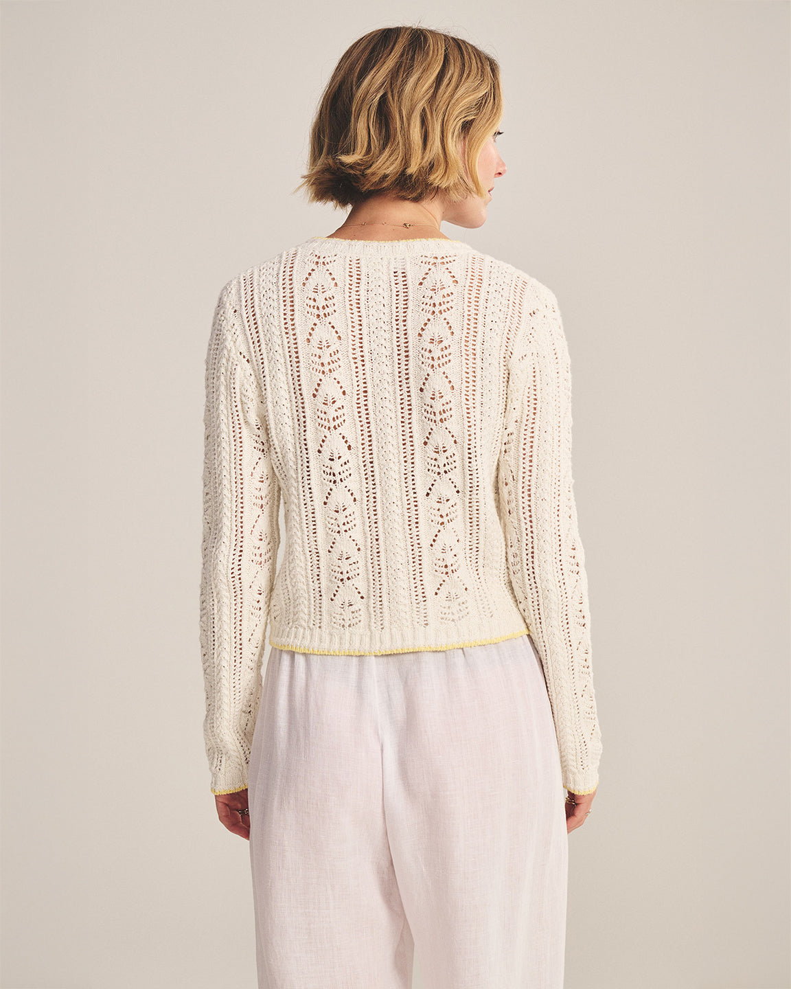 Woman wearing a cream-colored lace sweater and light pink pants against a plain background
