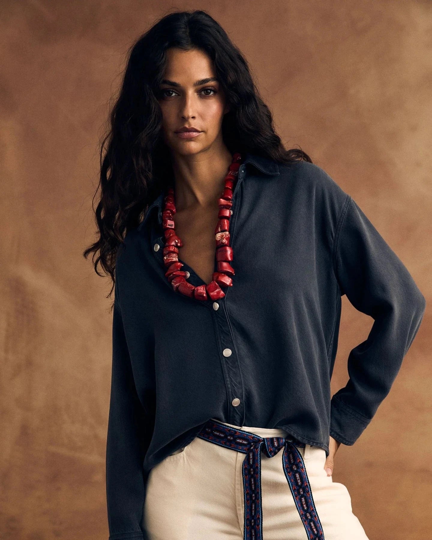 Woman wearing a washed black shirt and beige pants with a red necklace against a brown background