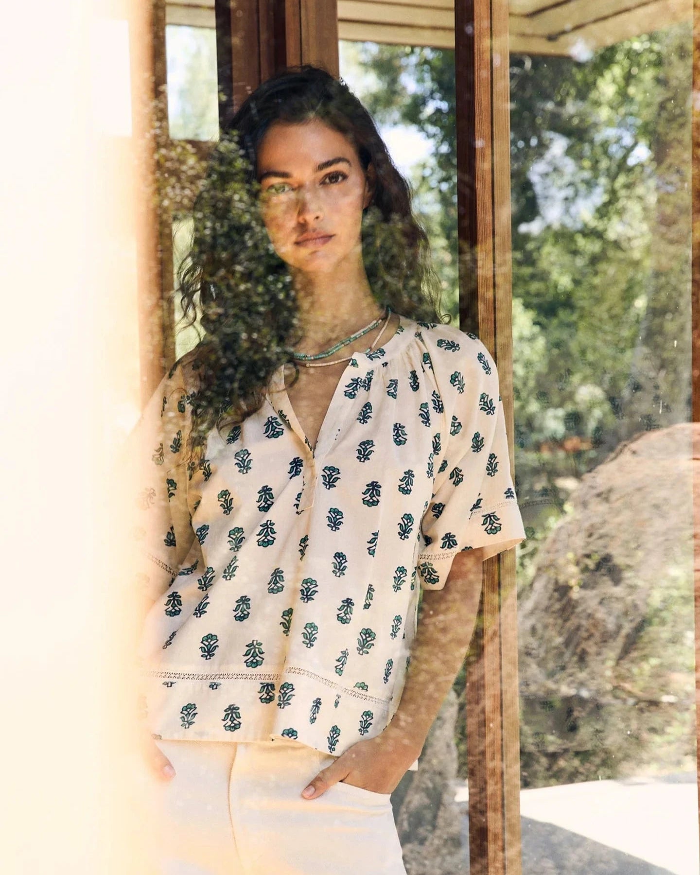 Woman wearing a patterned blouse and white pants standing in front of a glass door with a garden view.