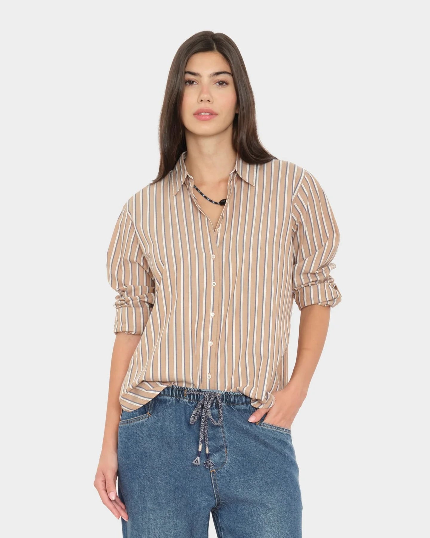 A woman modeling a beige and white vertically striped button-down shirt with rolled-up sleeves, paired with blue jeans, against a light gray background.