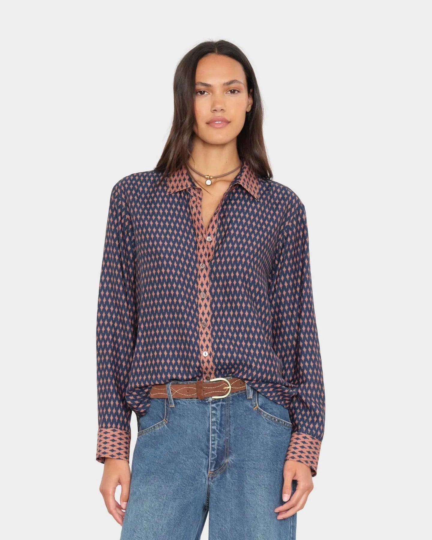 A woman modeling a long-sleeve button-up shirt with a small geometric pattern in navy and dusty pink, paired with blue jeans and a brown belt, against a light gray background.