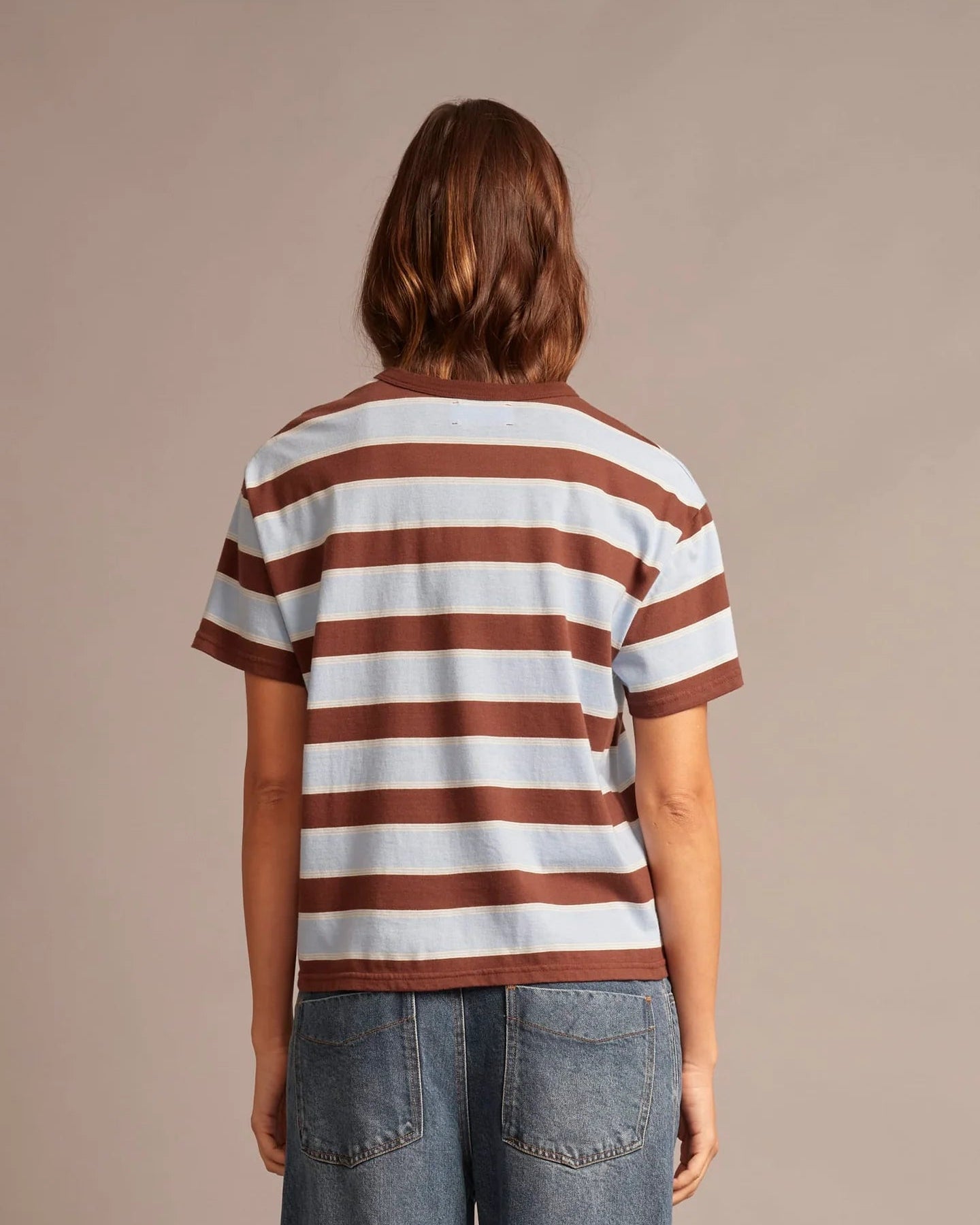A woman viewed from behind wearing a short-sleeve crewneck t-shirt with horizontal brown and off-white stripes, paired with blue jeans, against a solid beige studio background.