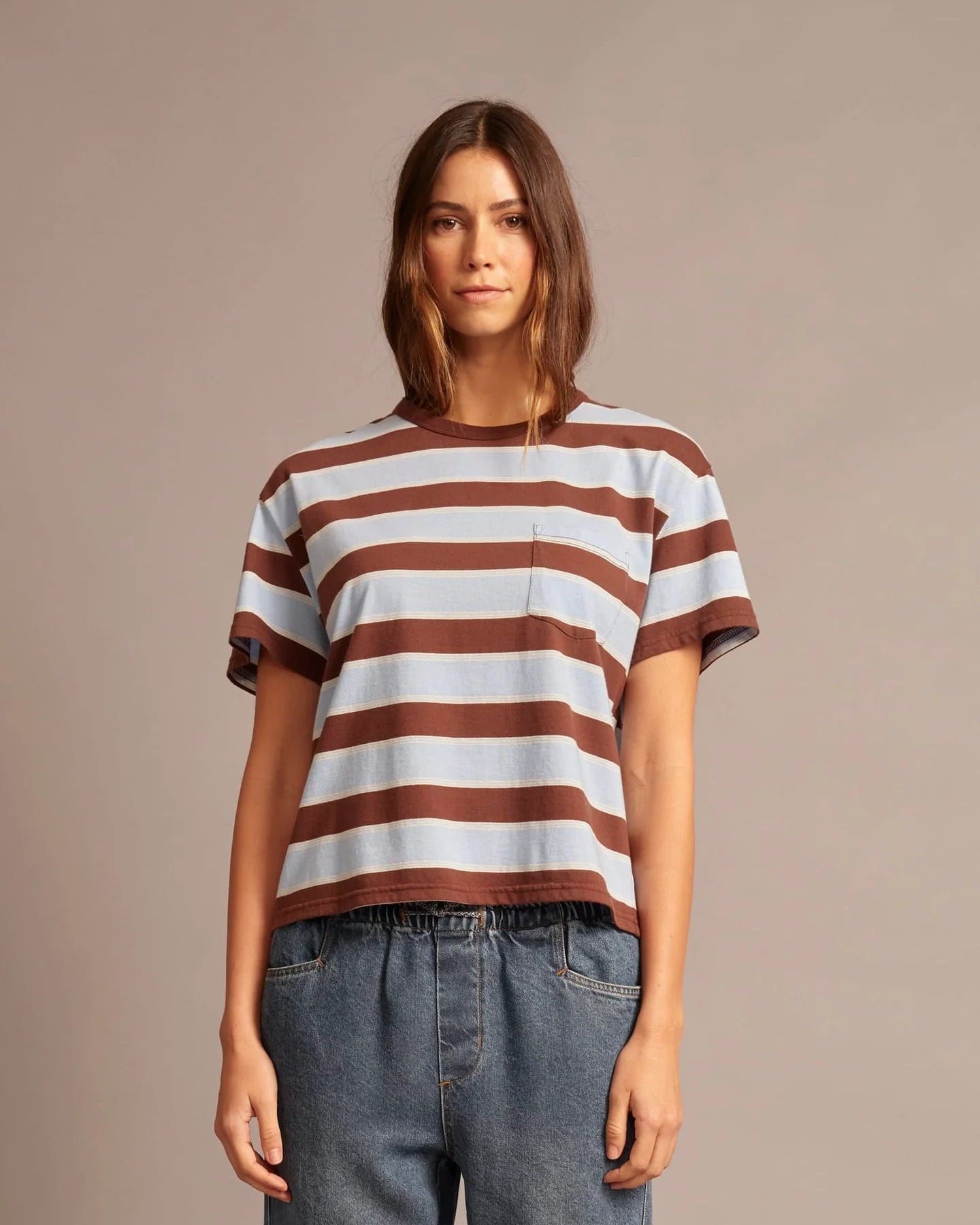 A woman wearing a brown and white horizontally striped short-sleeve t-shirt with blue jeans, standing against a solid warm gray studio background.