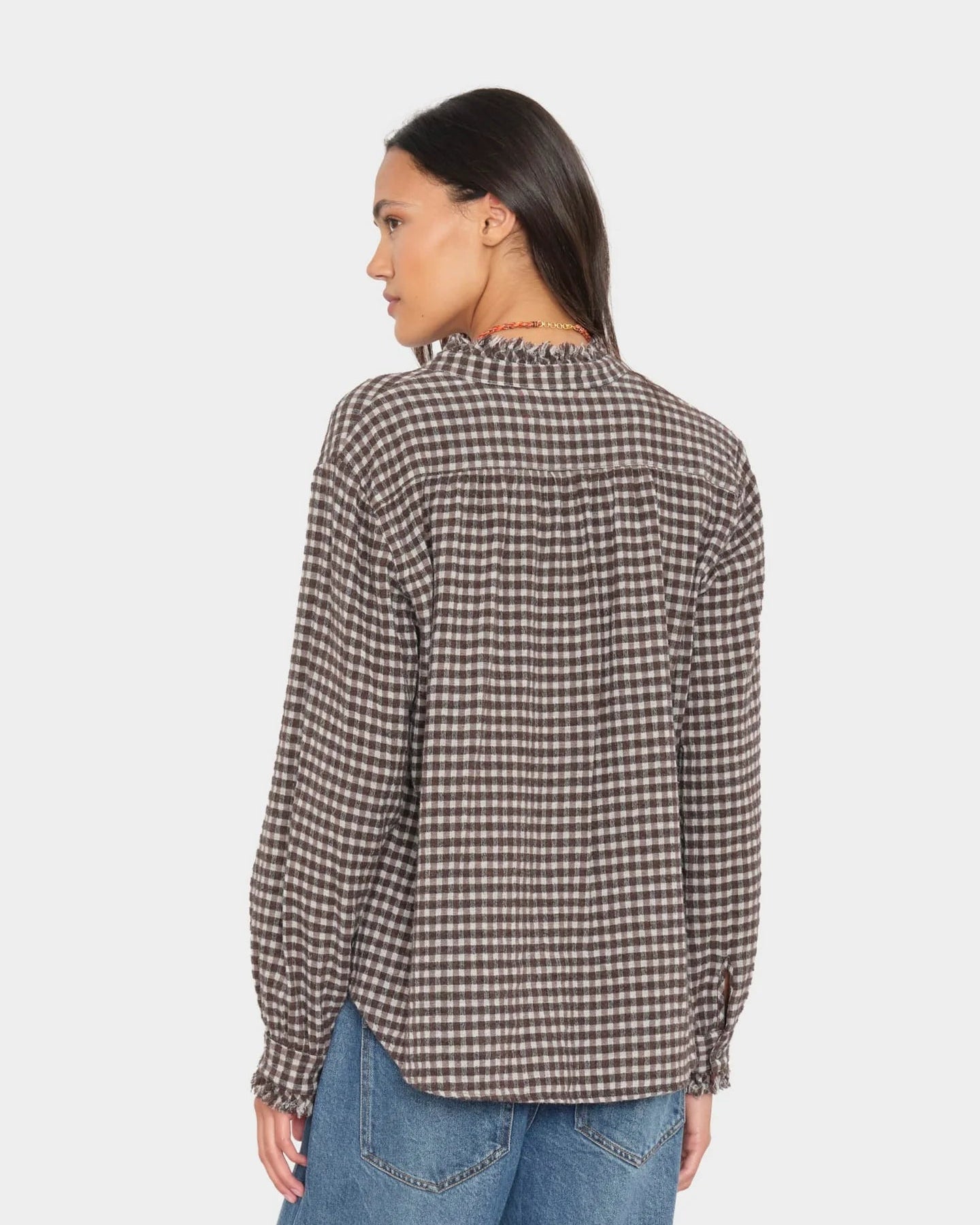 A woman shown from the back wearing a brown and beige checkered long-sleeve blouse with a ruffled high neckline, paired with blue jeans, against a light gray studio background.