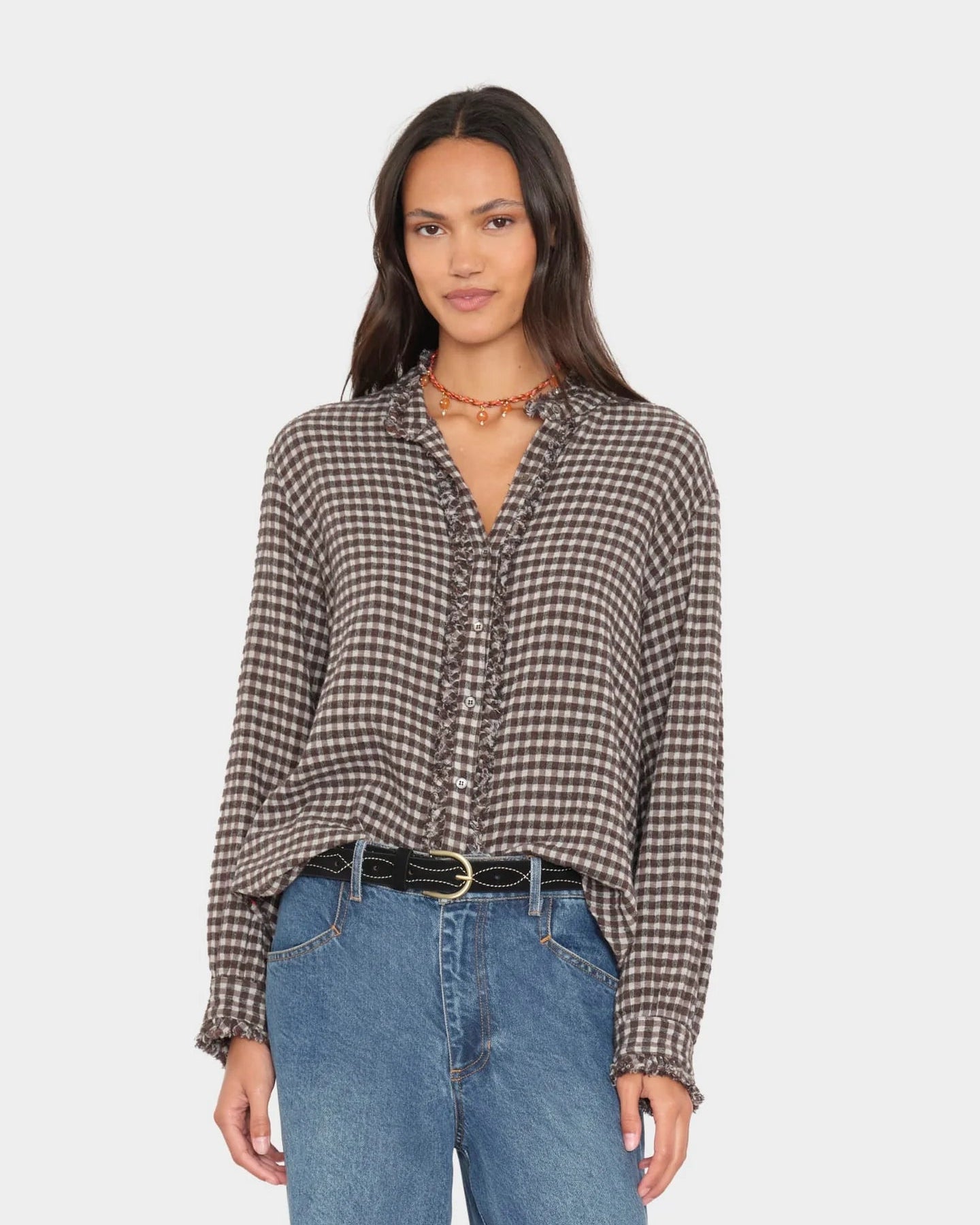 A woman modeling a brown and beige gingham long-sleeve blouse with a ruffled V-neckline, paired with blue jeans and a black belt, against a light gray studio background.