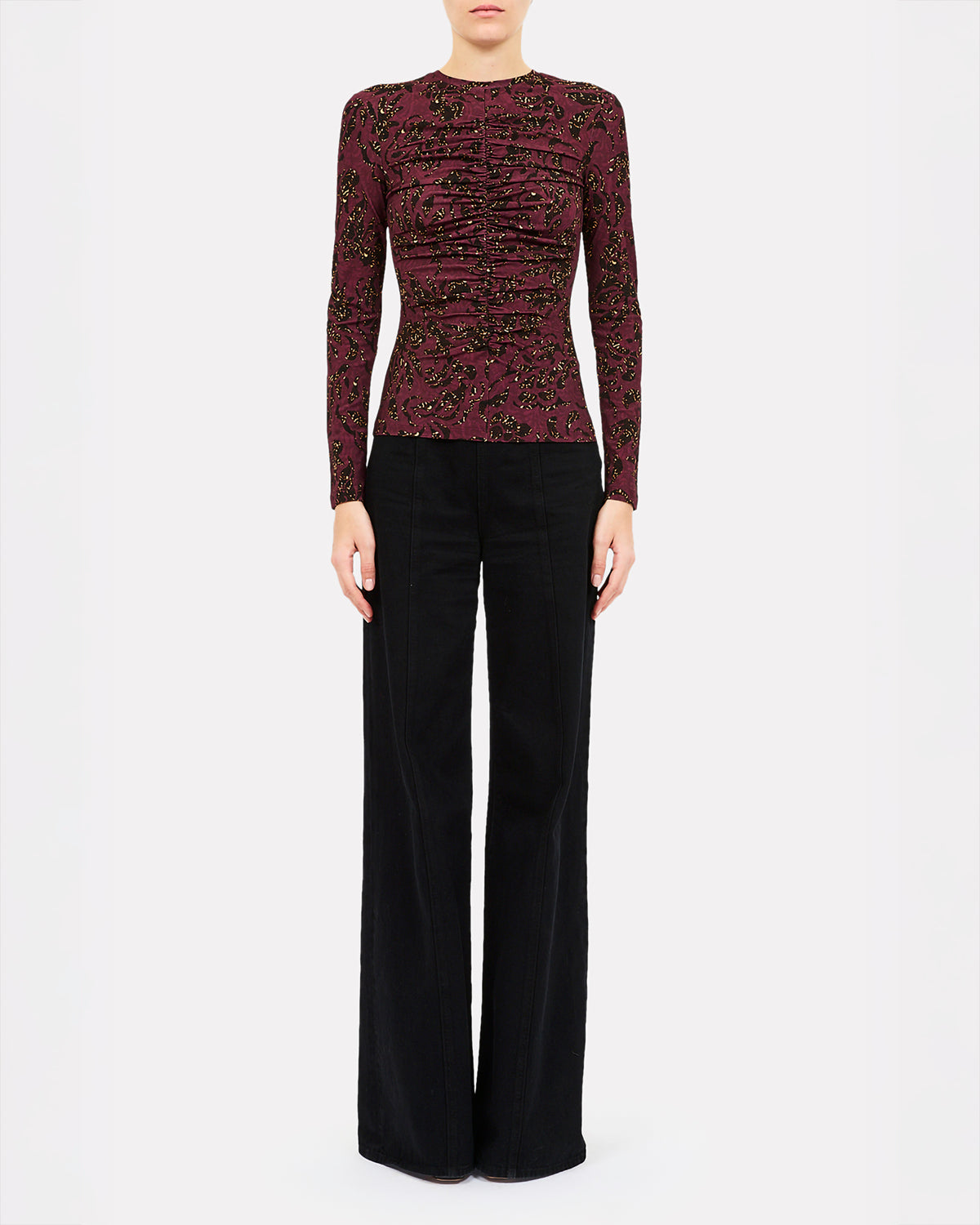 A model wearing a long-sleeve, ruched knit top in deep burgundy with black and gold floral embroidery, paired with black wide-leg trousers, shot against a plain light gray background.