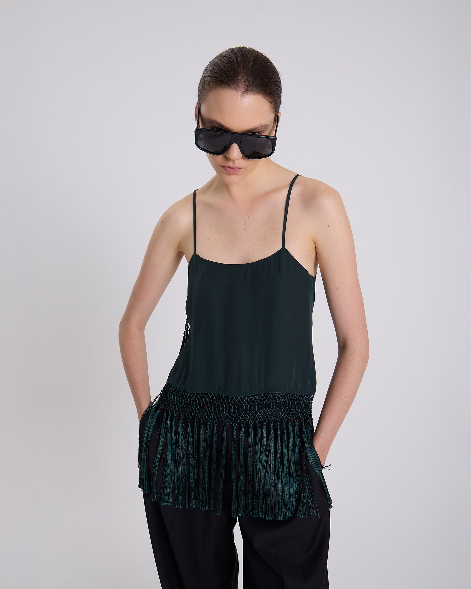 Woman wearing a dark green sleeveless top with fringe details on a plain background