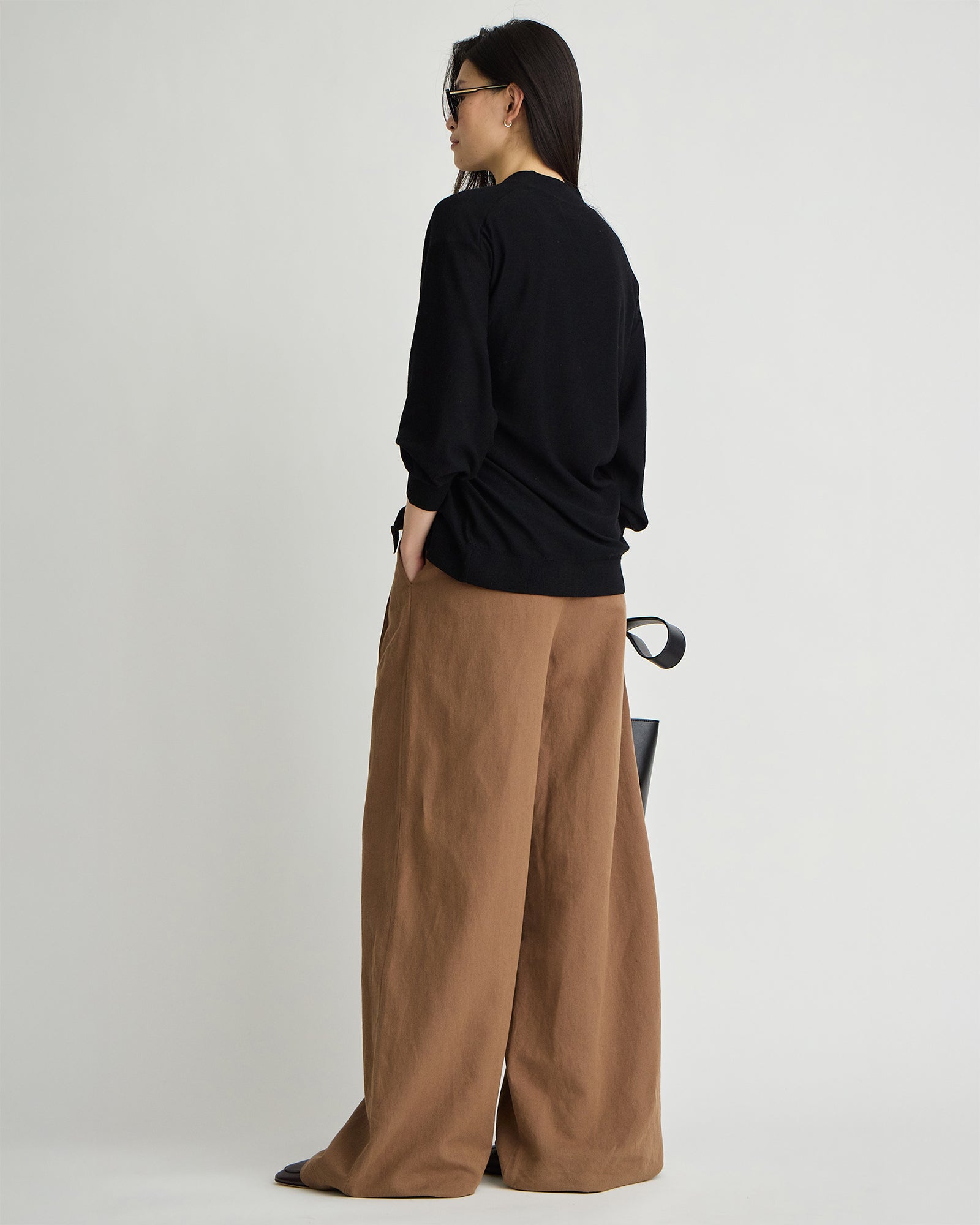 model wearing tobacco pleated pants and a black cardigan and sunglasses from the back