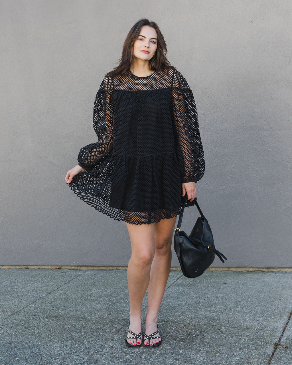 Woman wearing a black dress with sheer sleeves against a gray wall.