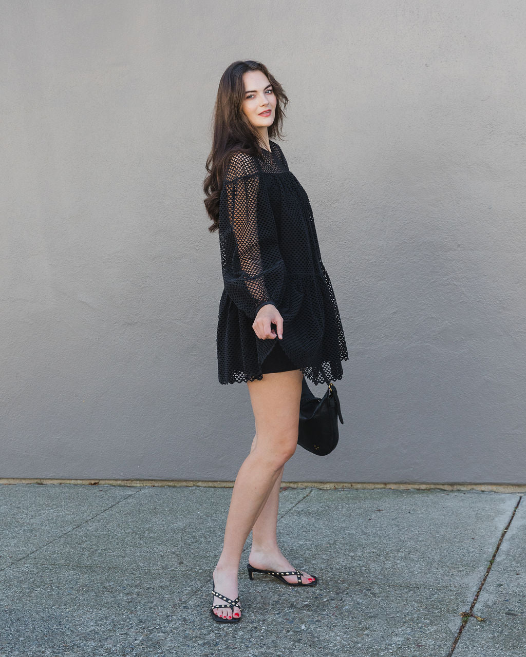 Woman wearing a black lace dress against a gray wall.