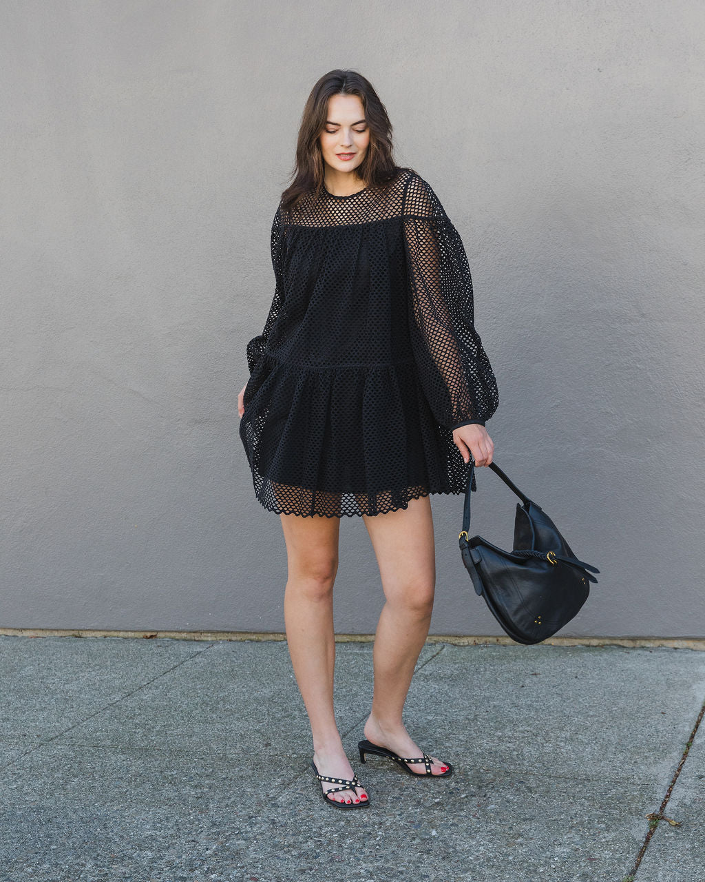 Woman wearing a black dress holding a black handbag against a gray wall.