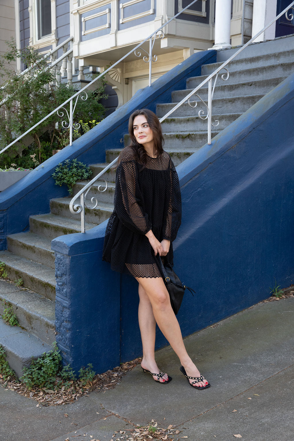 Woman in a black dress standing on a set of stairs with a blue wall and house in the background.