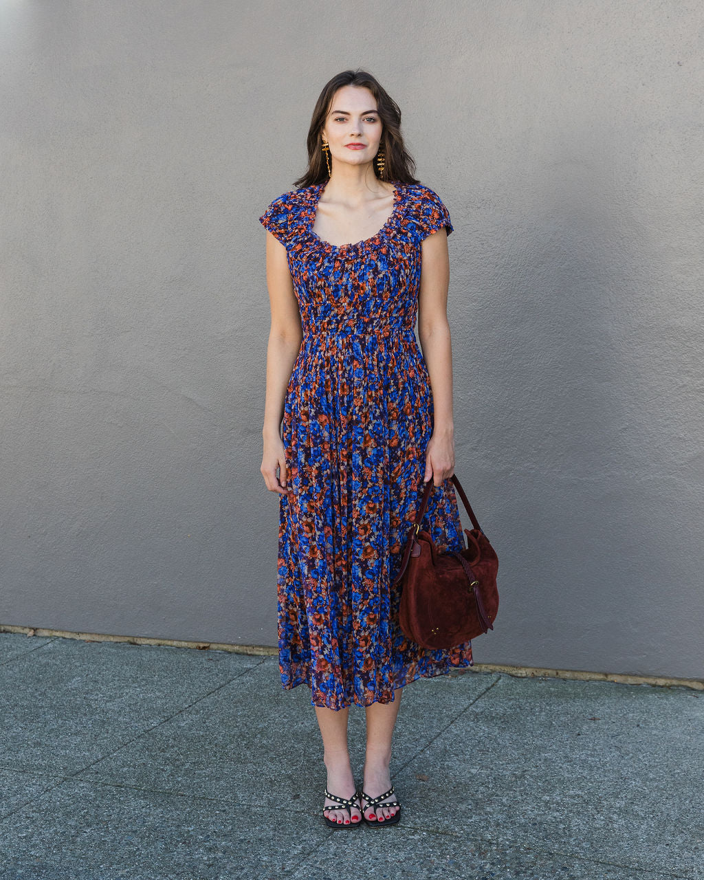 Woman wearing a blue floral dress holding a brown handbag against a gray background