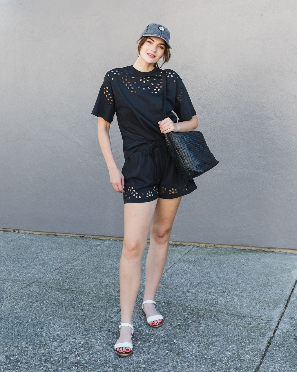 Woman wearing a black outfit with cut-out details, holding a black handbag against a gray wall.