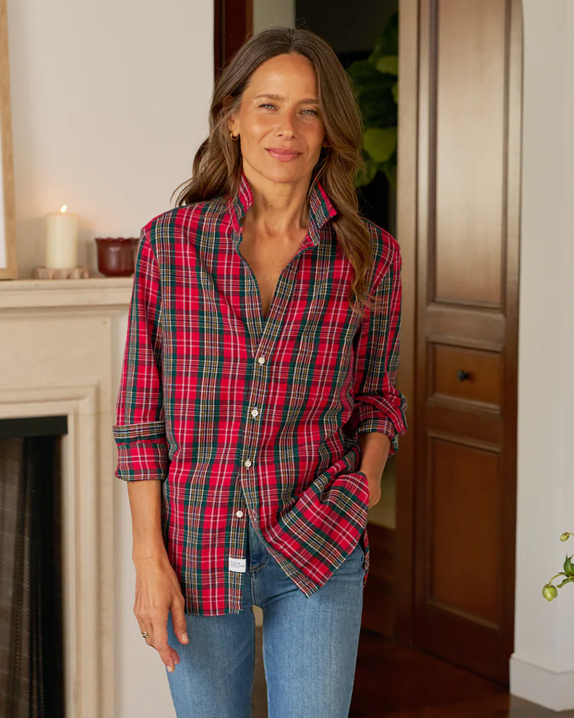 Woman wearing a red plaid shirt and blue jeans standing in a room with a fireplace.