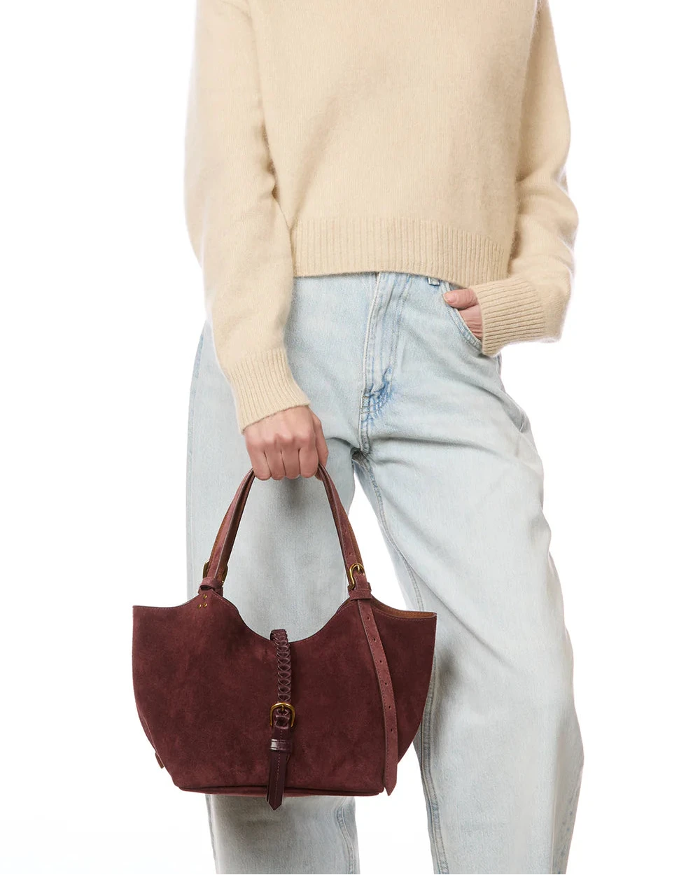 Person wearing a beige sweater and light blue jeans, holding a brown suede handbag on a white background