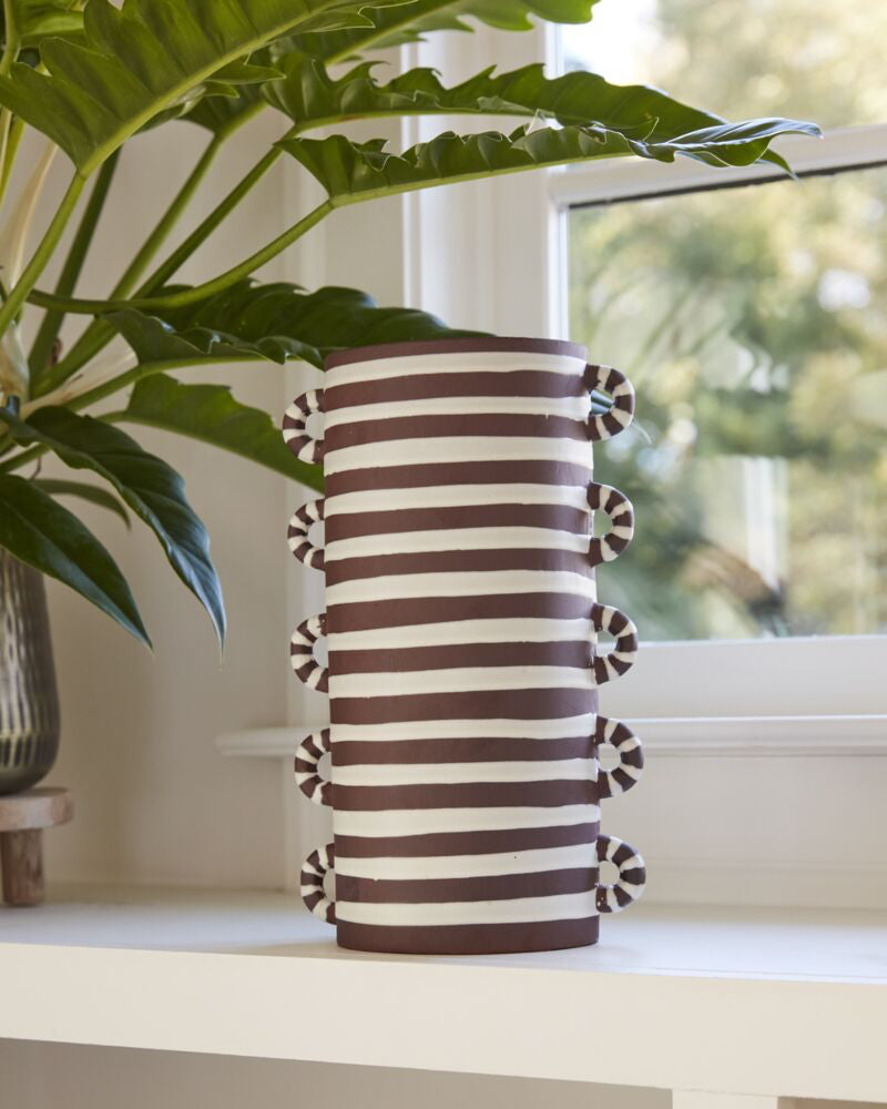 Striped vase with decorative elements on a shelf with plants in the background