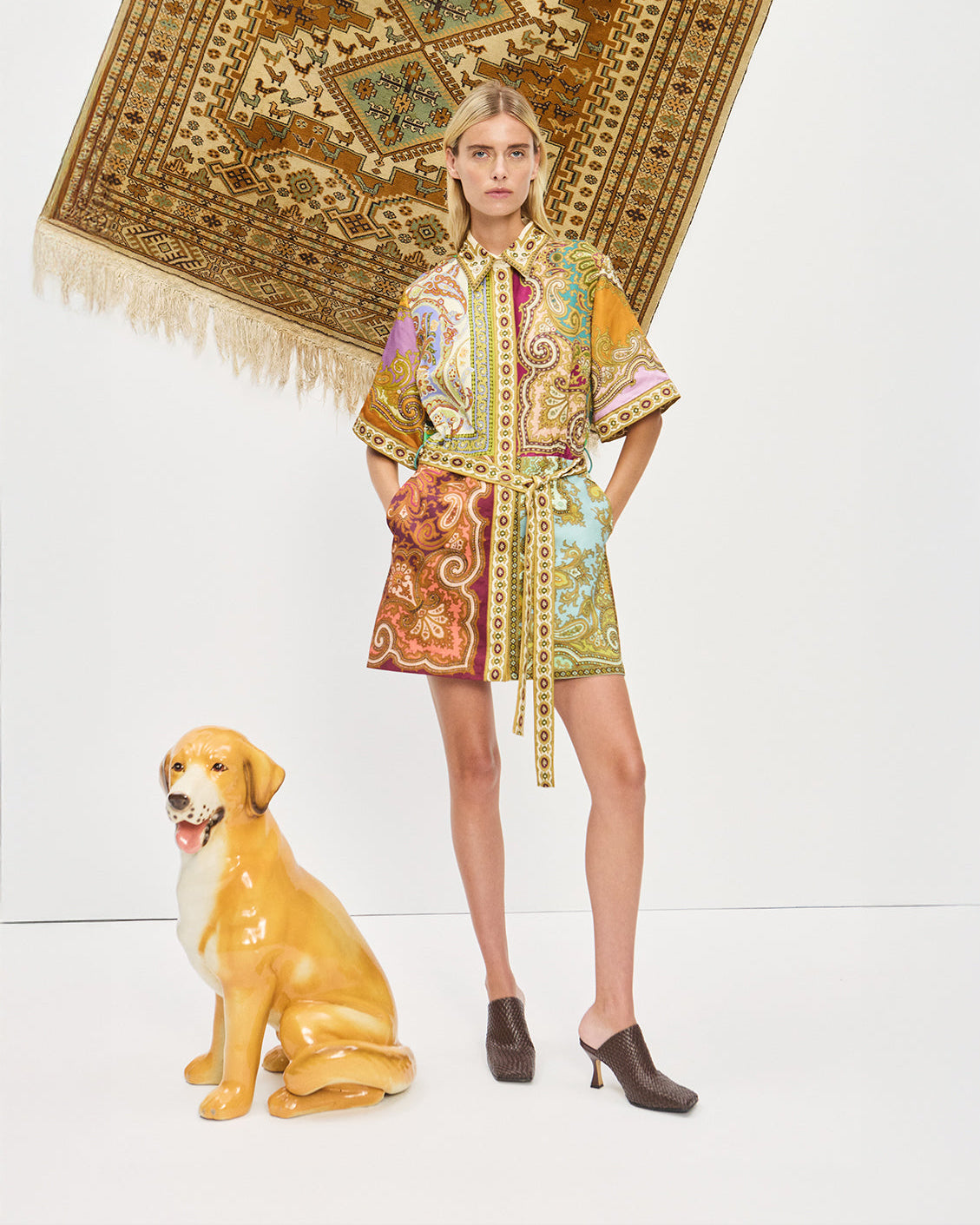Woman in a patterned outfit standing next to a dog with a decorative rug in the background