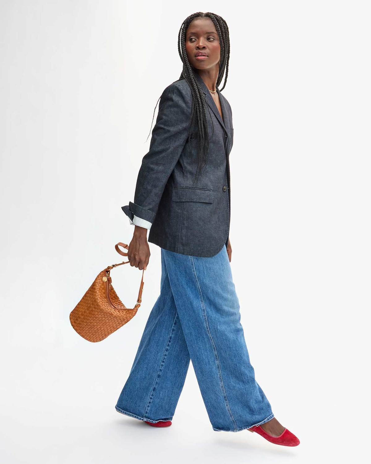 Woman wearing a gray blazer and blue jeans, holding a brown woven handbag on a white background