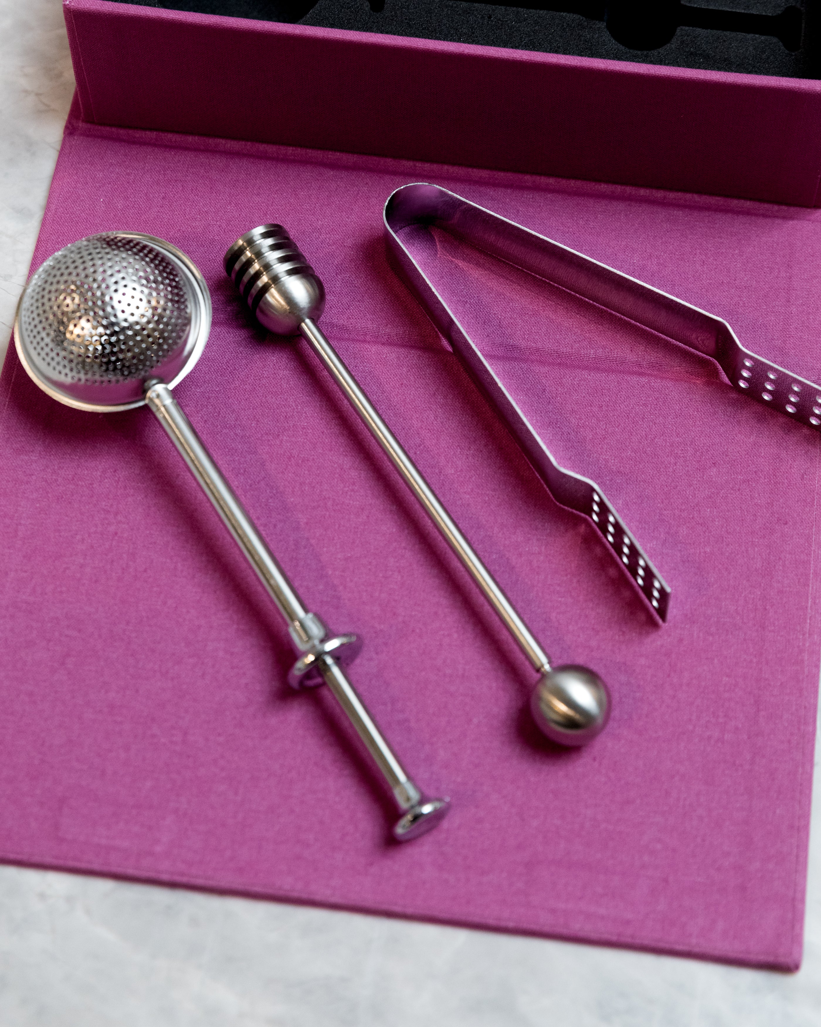 stainless steel tea strainer, tea tongs, and honey dipper on a purple background