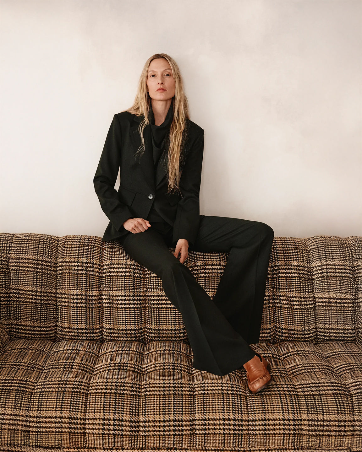 Woman in a dark green suit sitting on a patterned couch against a plain wall