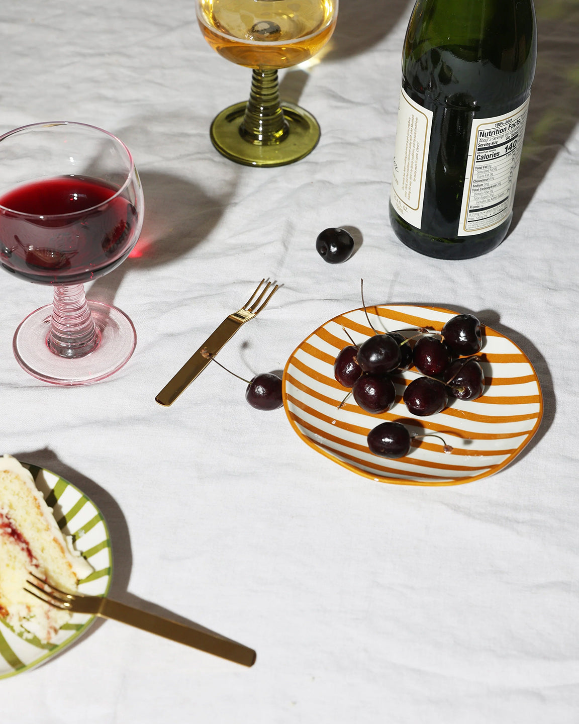 Table setting with wine glasses, cake, cherries, and a bottle on a white tablecloth.