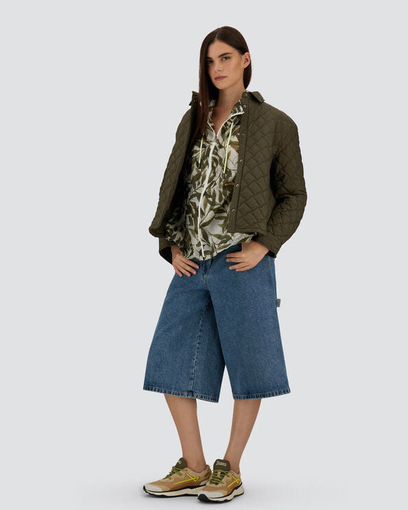 Woman wearing a green quilted jacket, camouflage shirt, and blue denim shorts on a white background