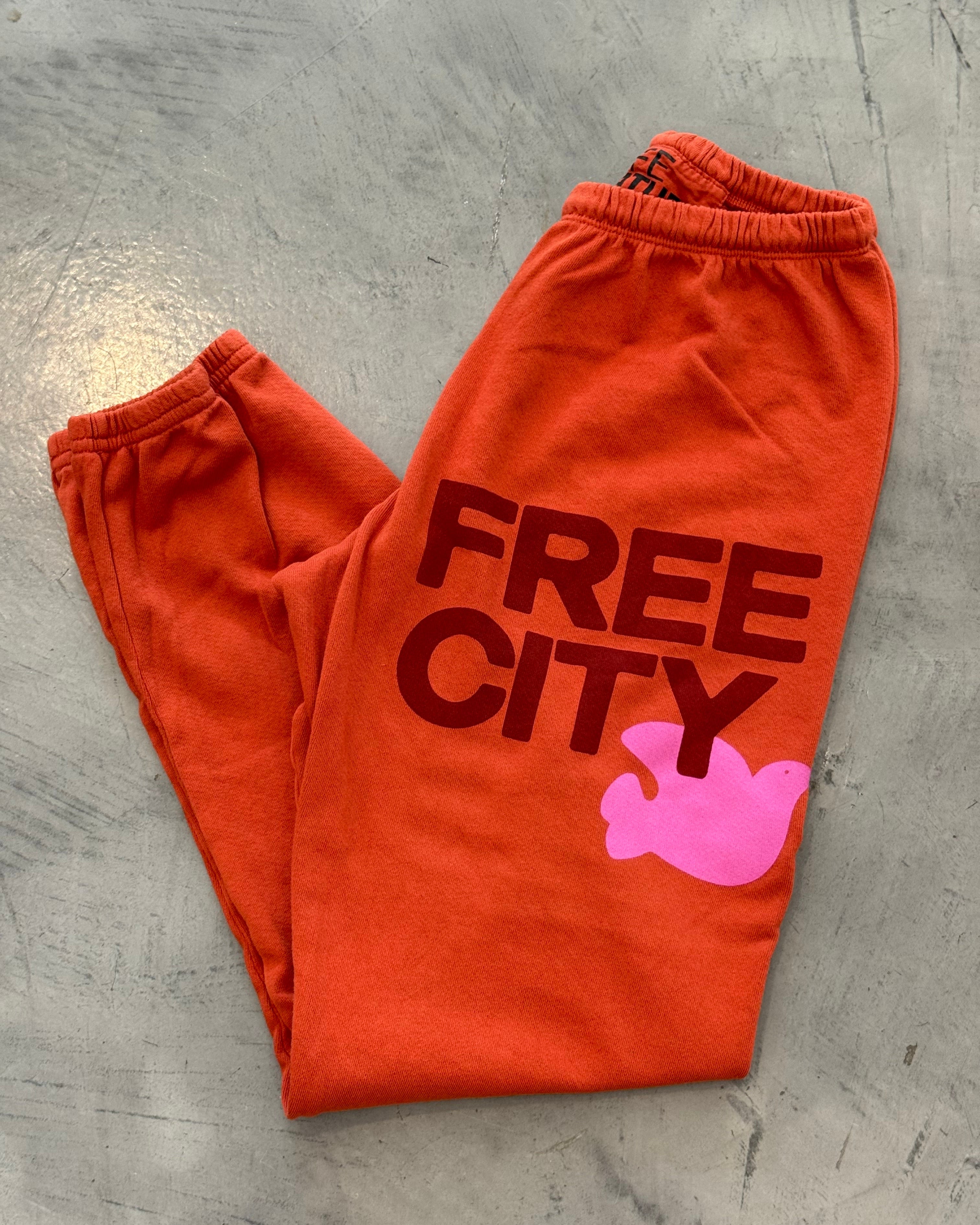 Orange sweatpants with 'FREE CITY' text and pink graphic on a gray surface