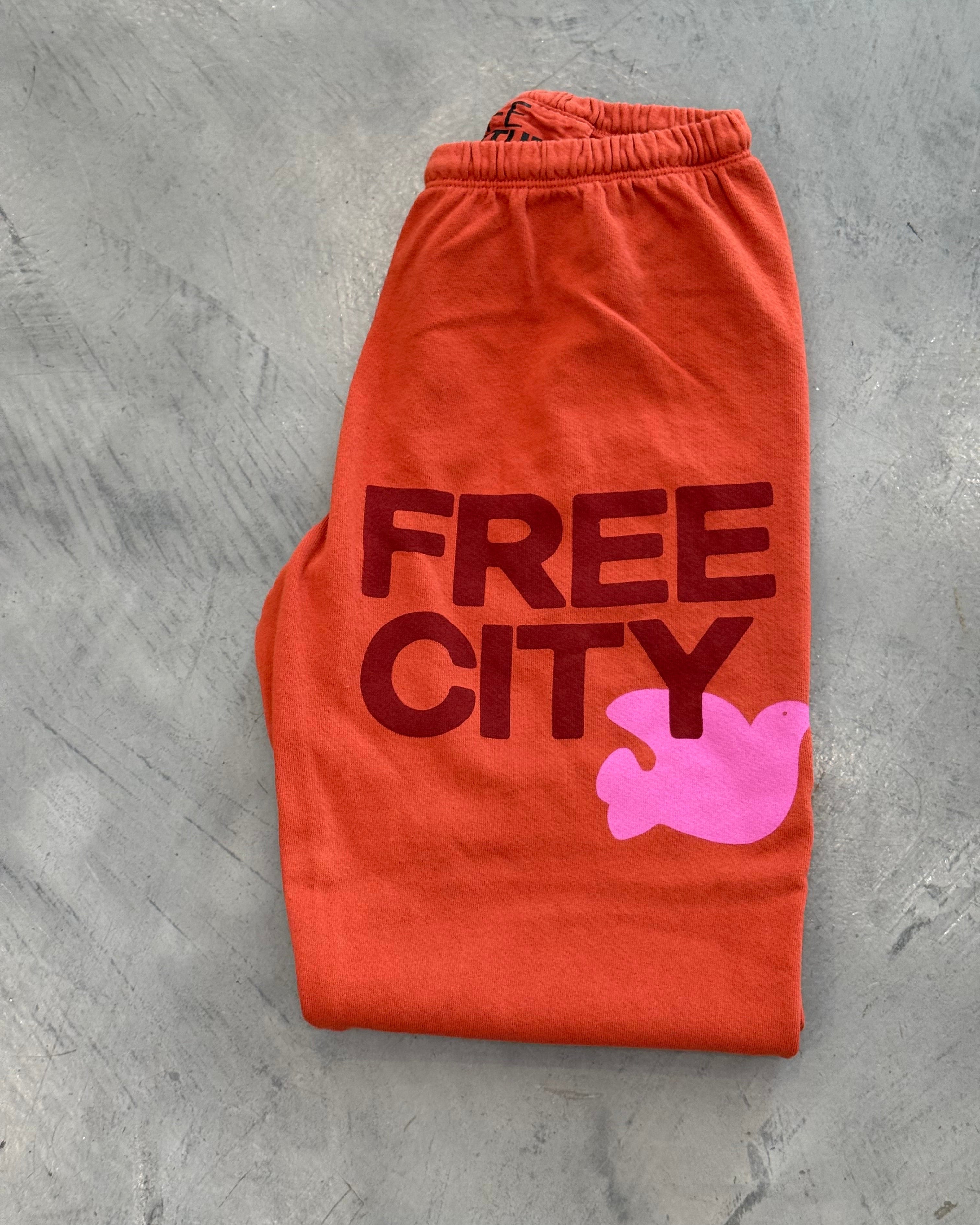 Orange sweatpants with 'Free City' text and pink graphic on a gray surface
