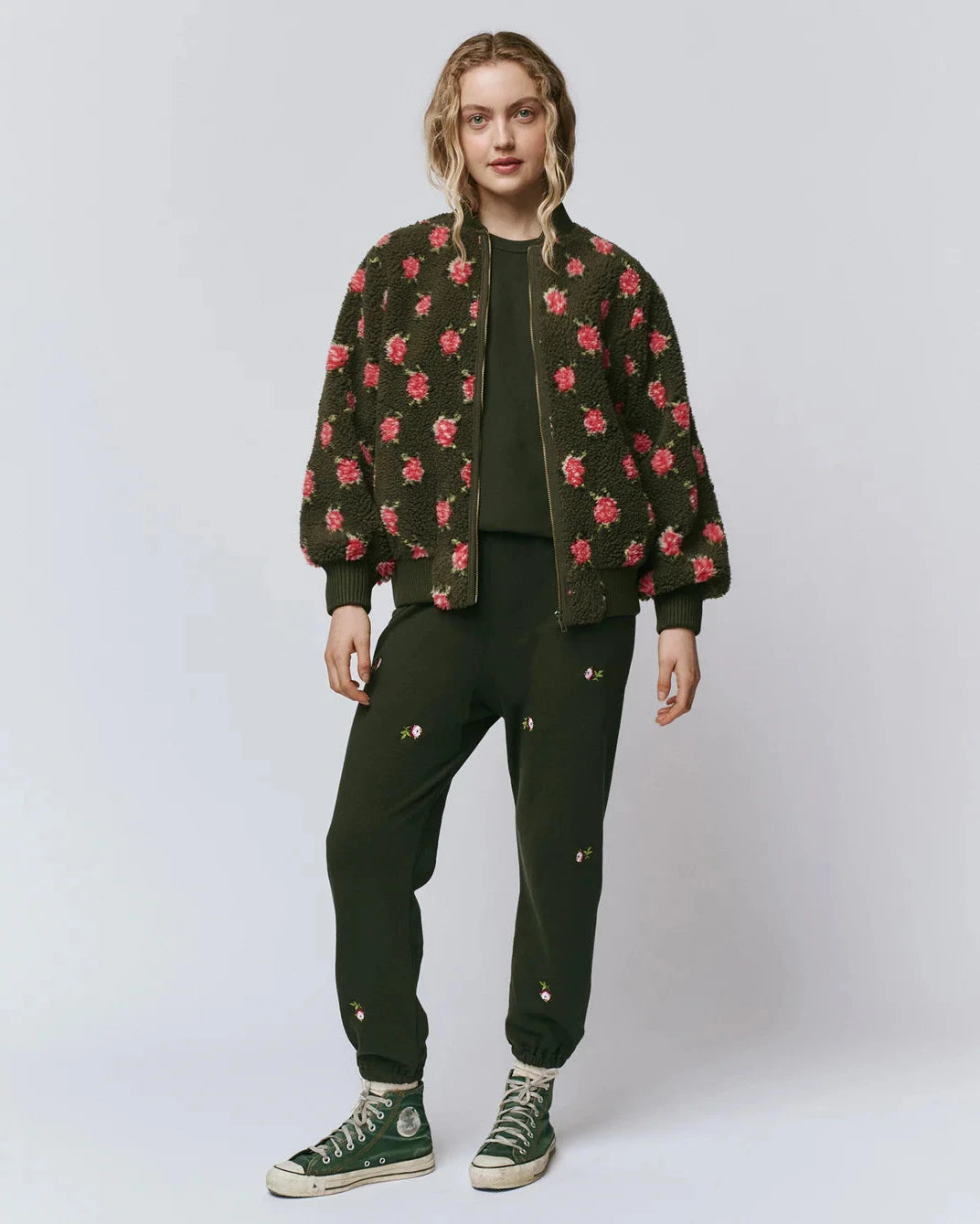 Person wearing a dark green jacket with red floral patterns, black pants, and green sneakers on a white background