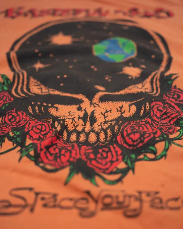 Close-up of an orange graphic t-shirt featuring a stylized skull with a starry night sky inside, red roses, and the text 'Space Your Face' in black script.