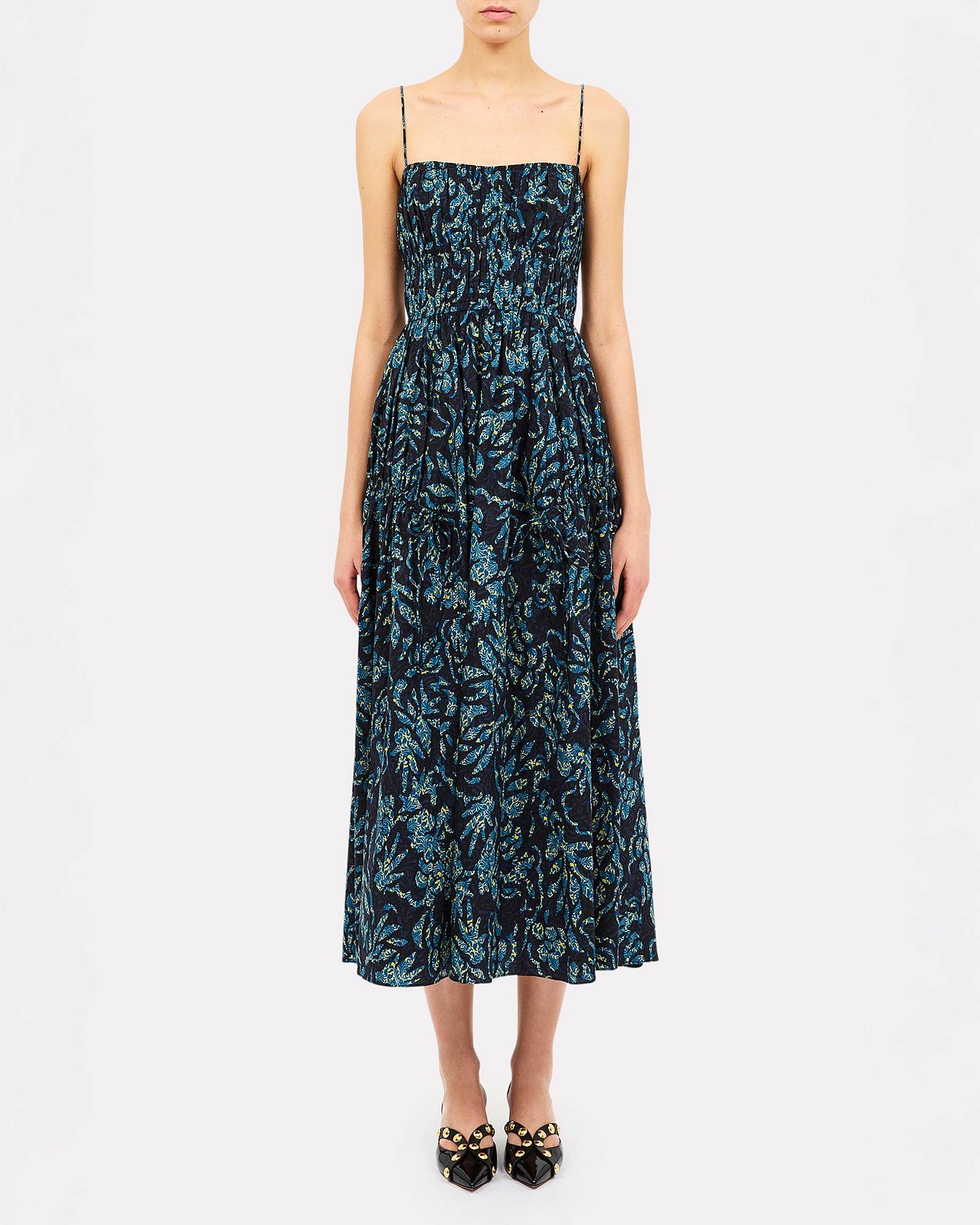 A woman modeling a sleeveless midi dress with thin straps and a dark blue and black floral pattern, paired with black pointed flats with gold embellishments, against a plain light gray background.