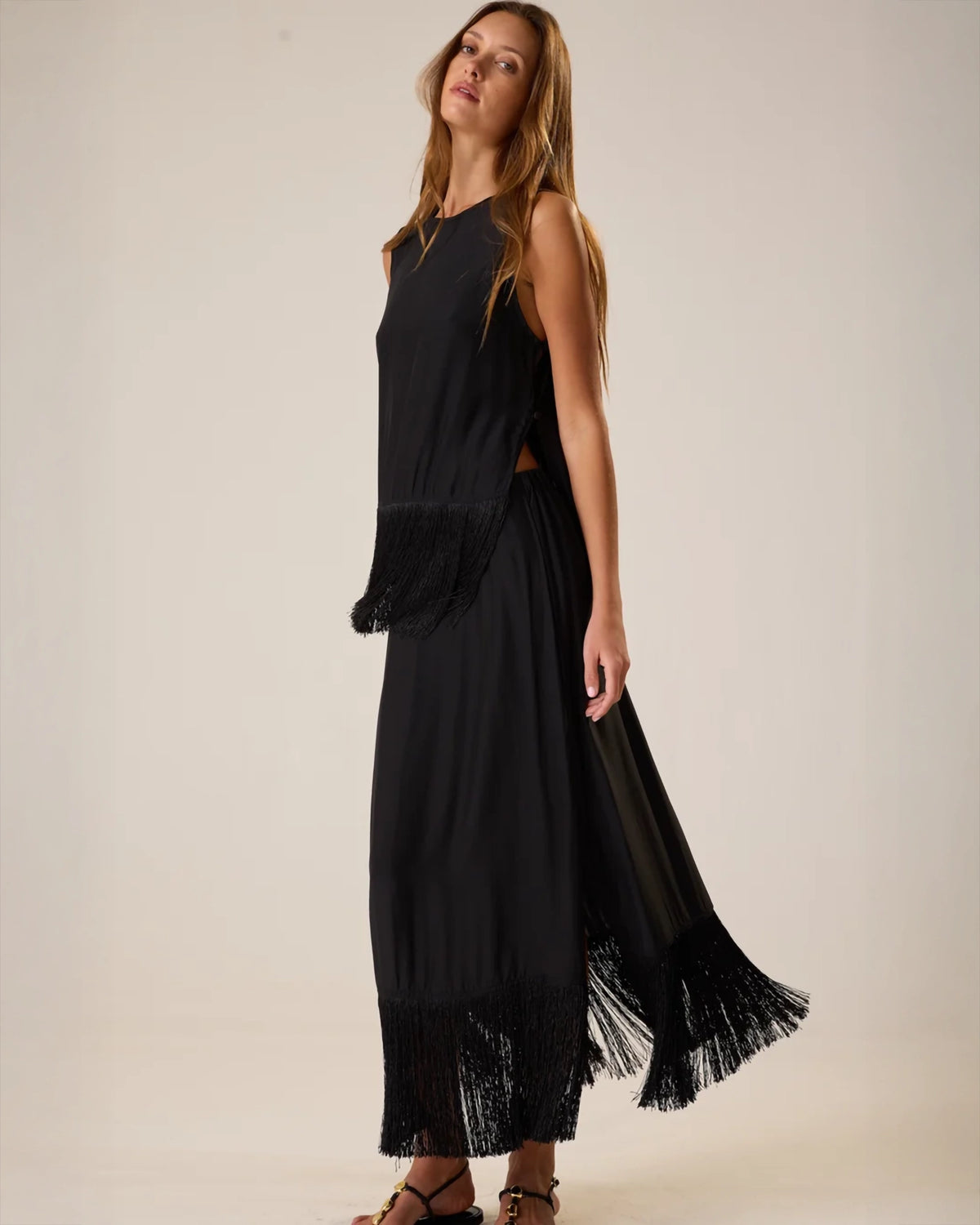 A woman modeling a sleeveless black maxi dress with fringe trim along the hem and a flowing side panel, photographed against a plain light beige studio background.