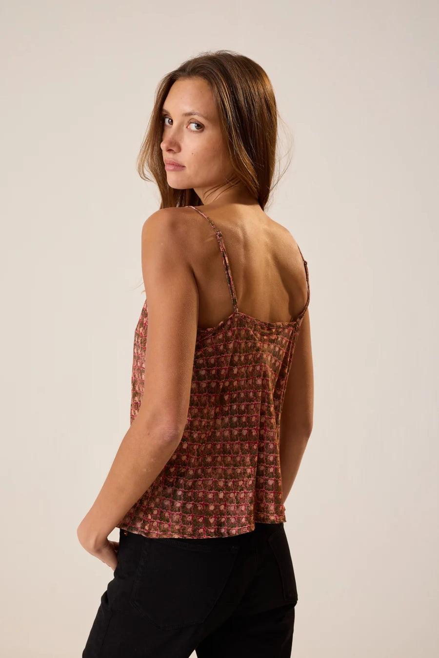 A woman shown from the back wearing a sleeveless patterned camisole top with thin straps and black pants, photographed against a plain light beige studio background.