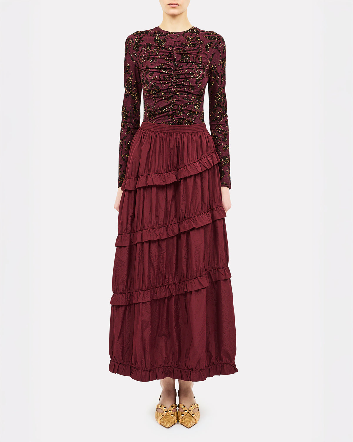 A model wearing a long-sleeved burgundy maxi dress with a heavily embellished lace bodice and a tiered ruffle skirt, paired with gold pointed flats, against a plain white studio background.