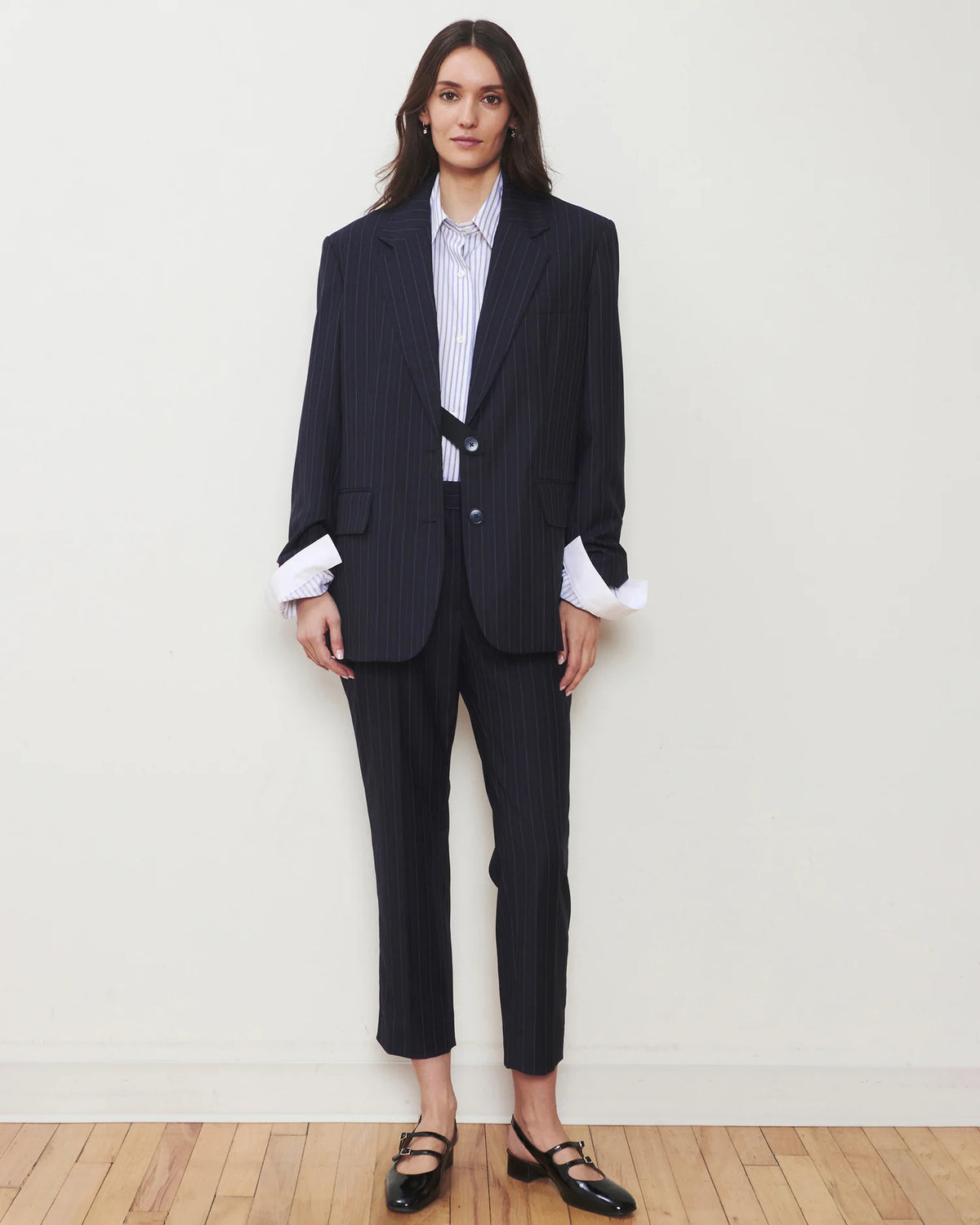 A woman stands against a plain off-white wall wearing a dark navy pinstripe suit with a white striped shirt and black Mary Jane flats.