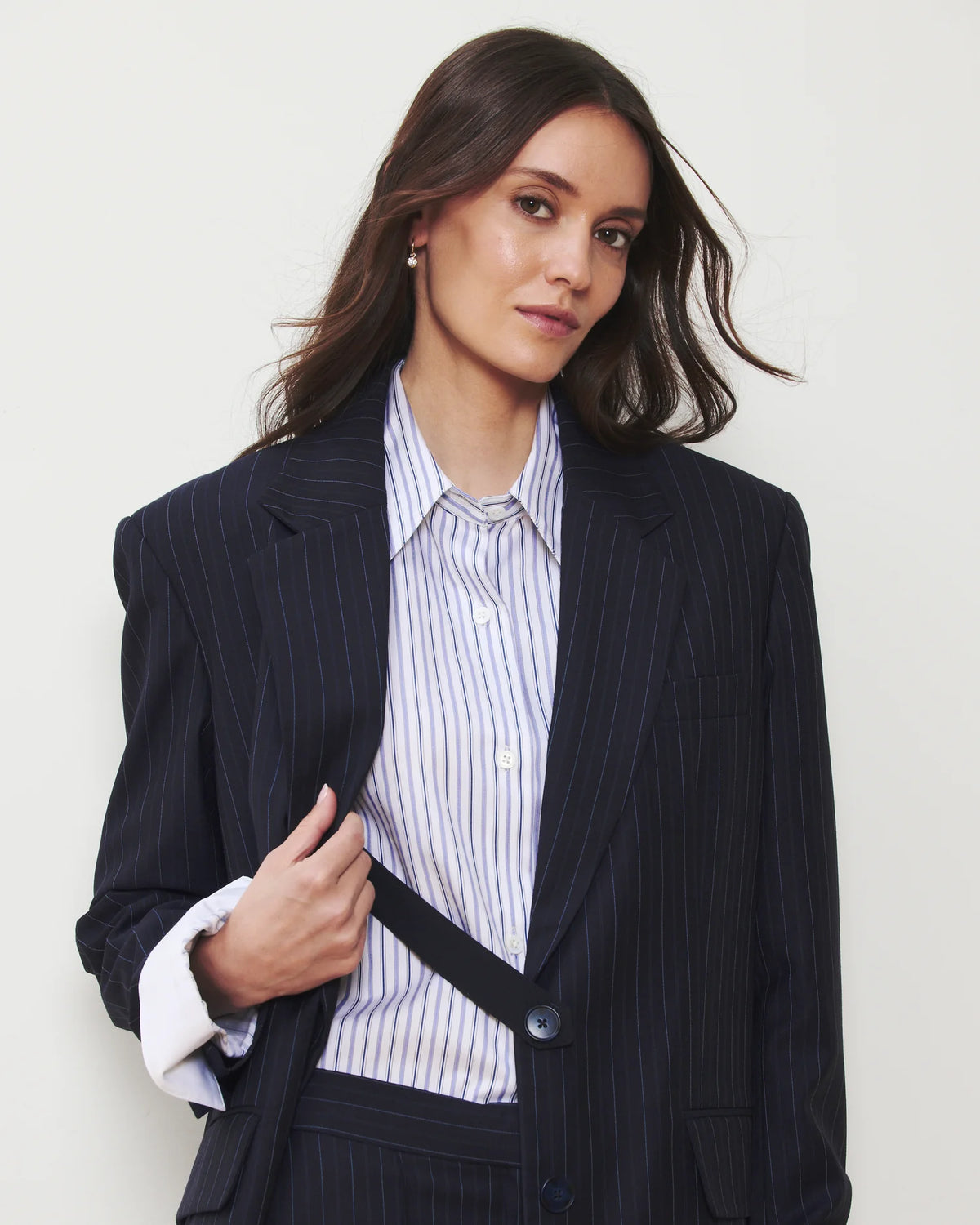 A woman wearing a dark navy pinstripe blazer over a white and navy striped button-down shirt, posed against a light off-white background.