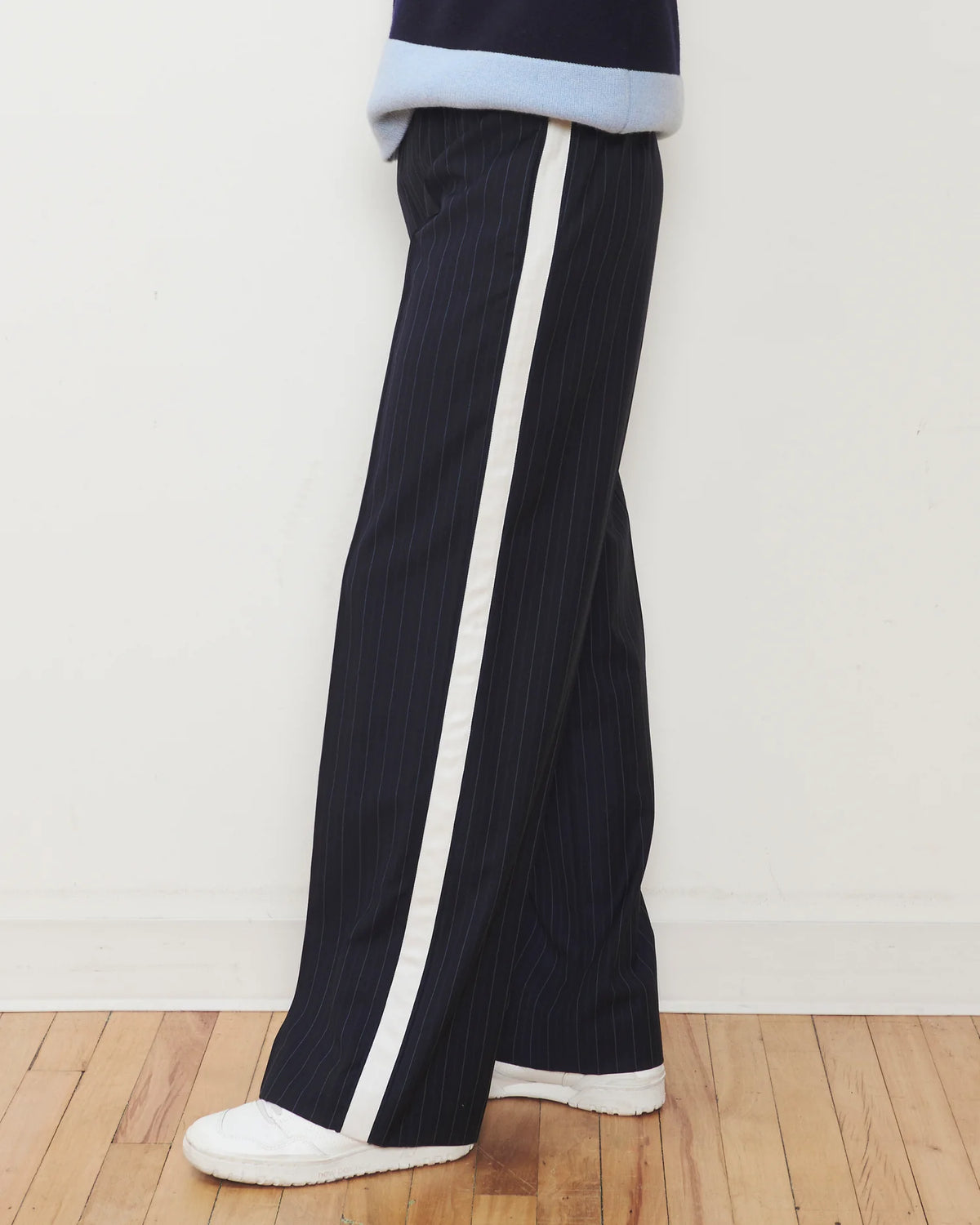 A person wearing dark navy pinstriped wide-leg trousers with a prominent white side stripe, paired with white sneakers and a light blue and navy top, standing on a wooden floor against a white wall.
