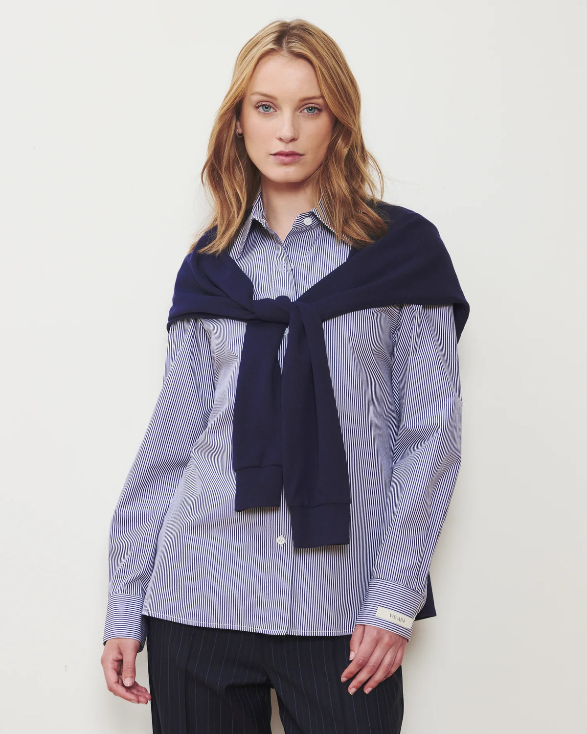A woman modeling a light blue and white vertically striped button-down shirt, styled with a dark navy knit sweater draped over her shoulders and dark pinstriped trousers, against a plain off-white background.