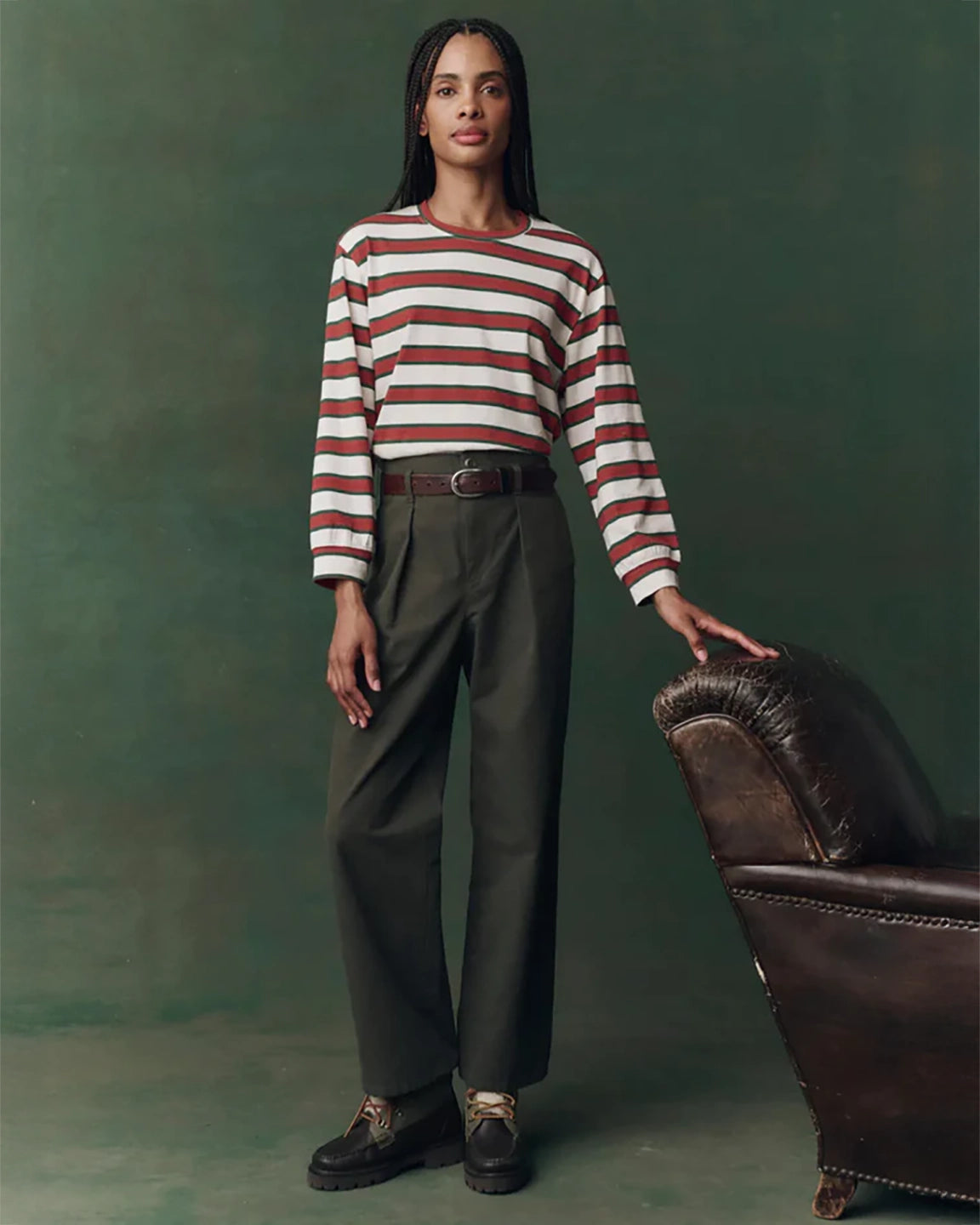Woman in a striped sweater and pants standing next to a leather chair against a green background