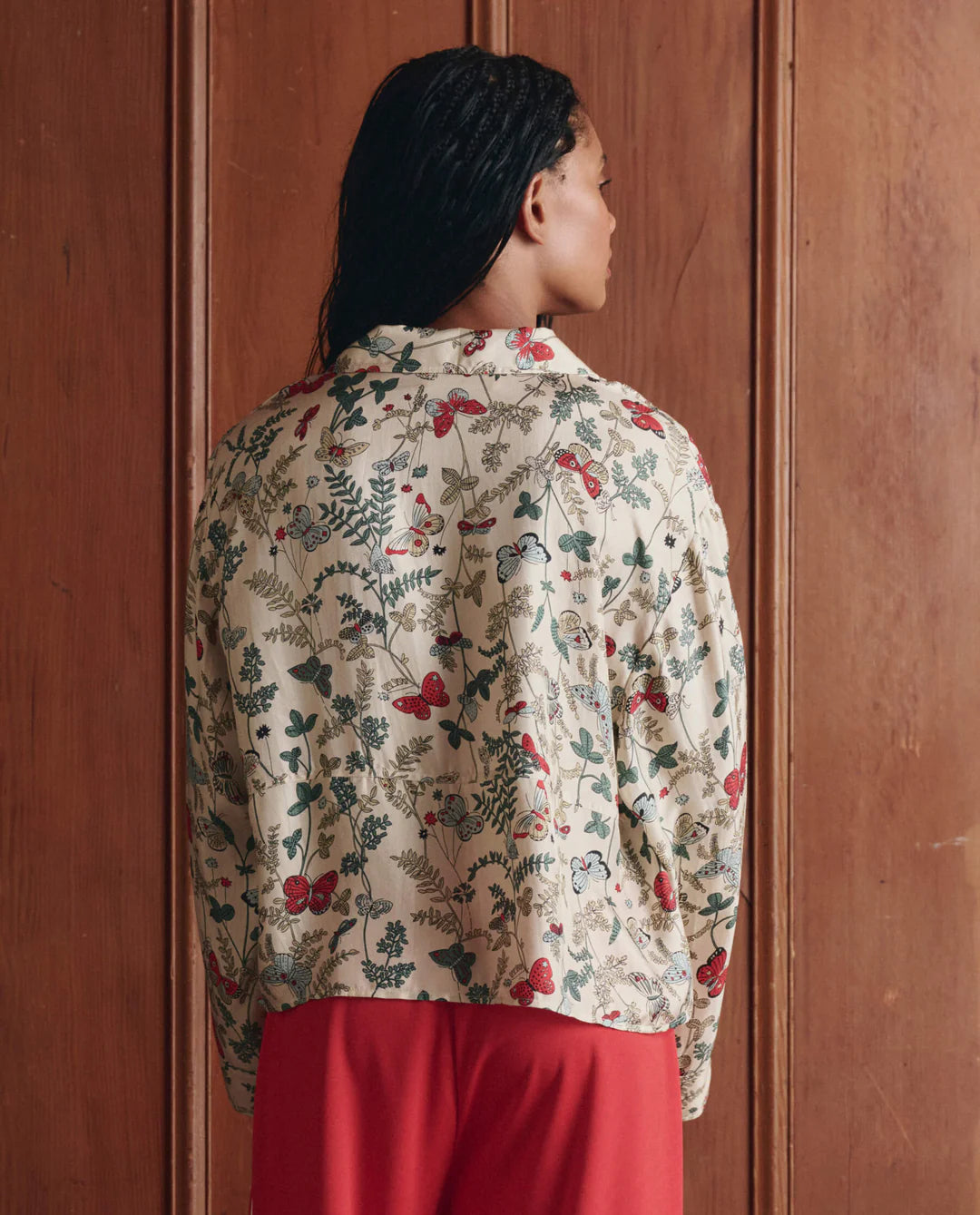 Woman wearing a floral blouse and red pants against a wooden panel background
