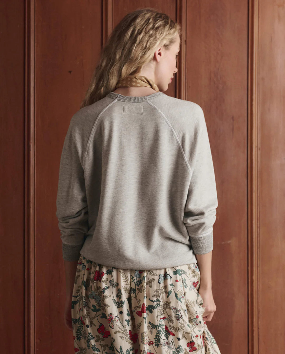 Person wearing a gray sweatshirt and floral skirt standing against a wooden door.