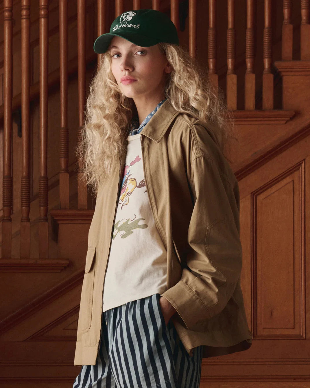 Person wearing a beige jacket, white graphic t-shirt, and striped pants standing in front of wooden stairs.