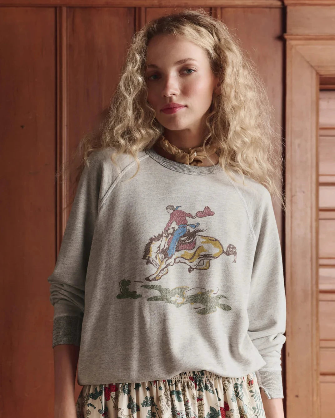 Woman wearing a gray sweatshirt with a graphic design in front of wooden paneling.